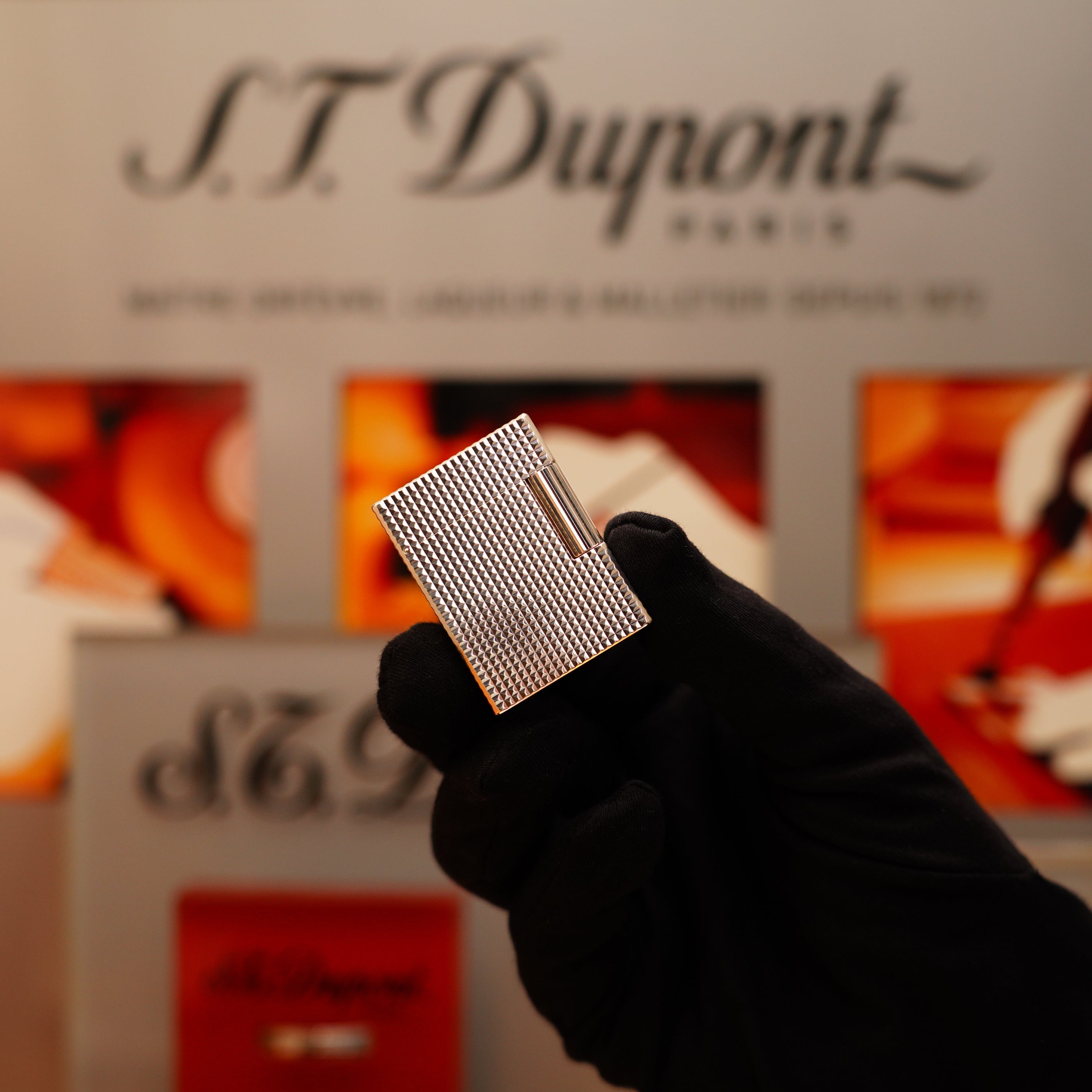 A gloved hand holds a Vintage 1996 S.T. Dupont Ligne 1 Diamond Head Pattern lighter with a heavy silver finish in front of a blurred display featuring "S.T. Dupont Paris.