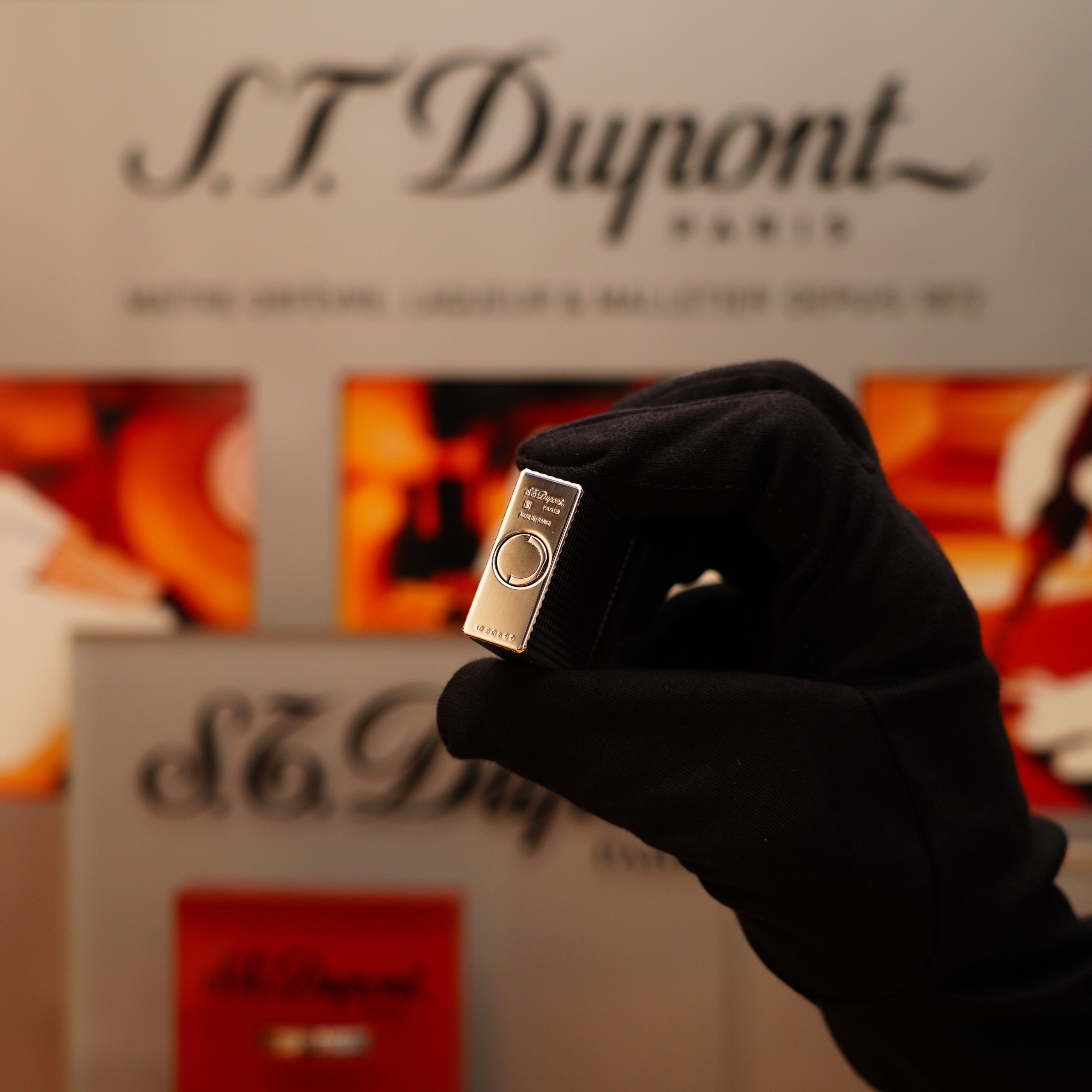 A gloved hand holds a Vintage 1996 S.T. Dupont Ligne 1 Diamond Head Pattern small Heavy Silver Finish Lighter in front of a blurred brand display.
