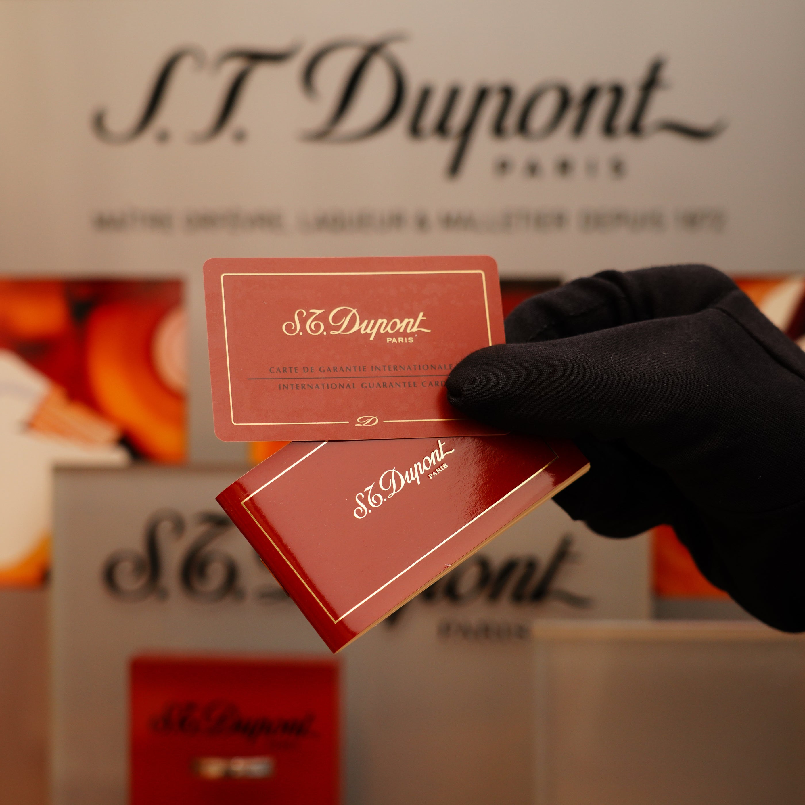A gloved hand holds warranty cards from S.T. Dupont in front of a blurred display featuring a Vintage 1996 S.T. Dupont Ligne 1 Diamond Head Pattern small Heavy Silver Finish Lighter.