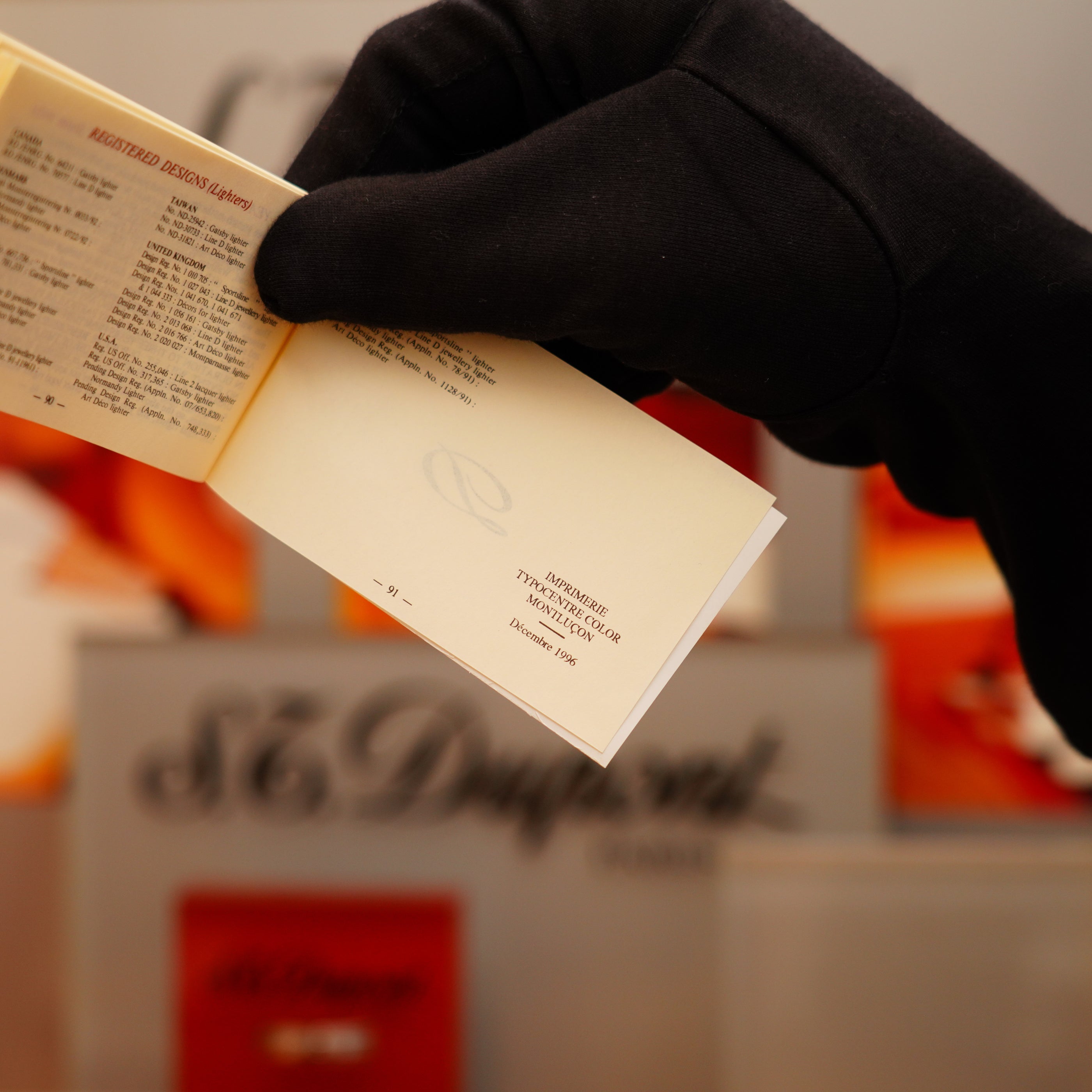 A person wearing black gloves holds a small booklet with text in front of blurred red and gray packaging for the S.T. Dupont Vintage 1996 Ligne 1 Diamond Head Pattern Lighter, featuring a heavy silver finish.