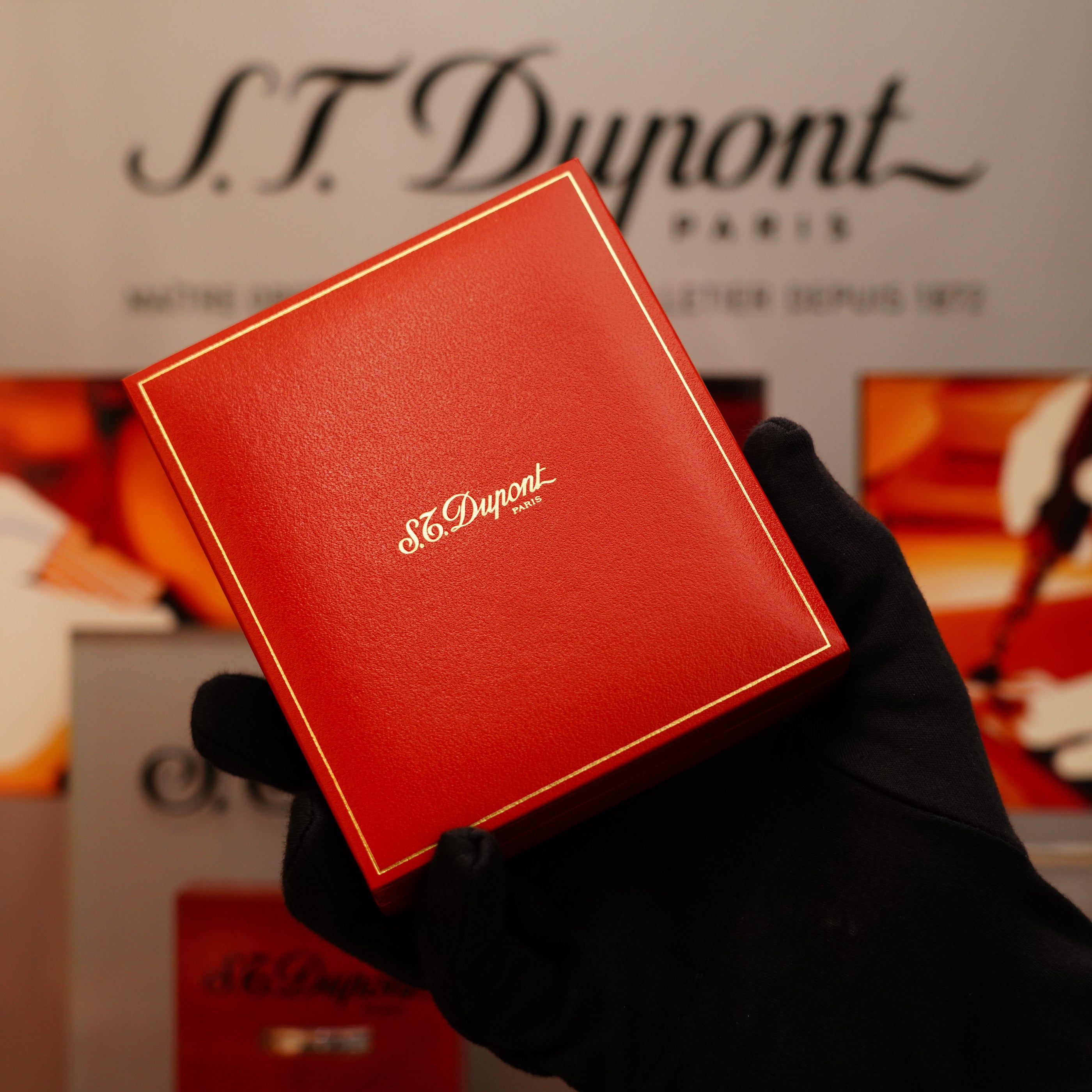 A gloved hand holds a red box for the Vintage 1996 S.T. Dupont Ligne 1 Diamond Head Pattern small Heavy Silver Finish Lighter, with a blurred S.T. Dupont branding in the background.