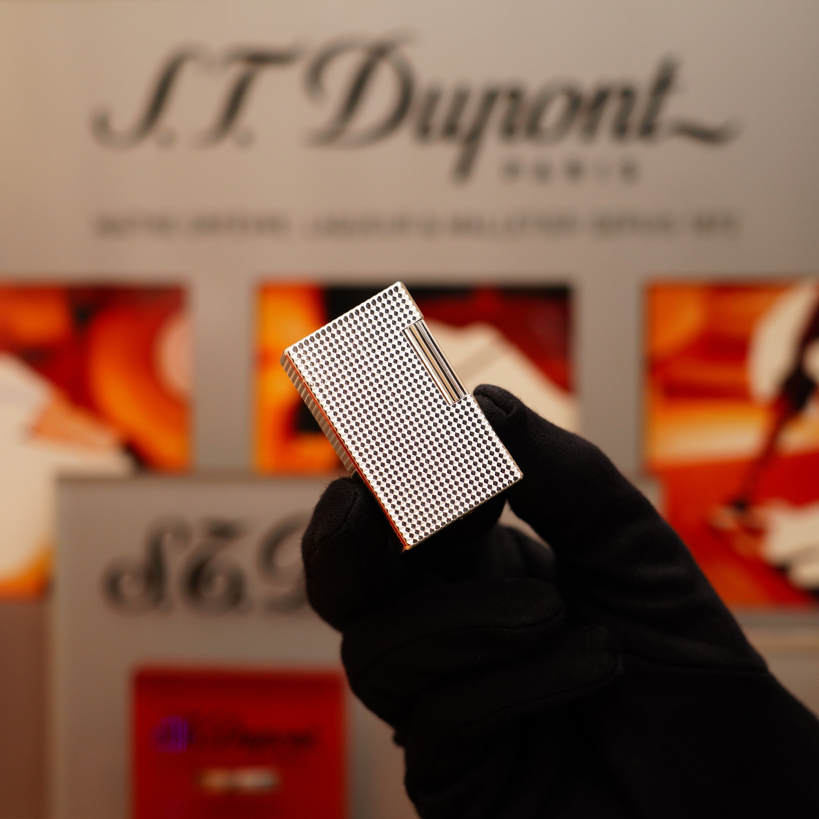 A gloved hand holds a Vintage 1996 S.T. Dupont Ligne 1 Diamond Head Pattern Large Heavy Silver Finish Lighter against a blurred background with the S.T. Dupont logo and colorful designs.