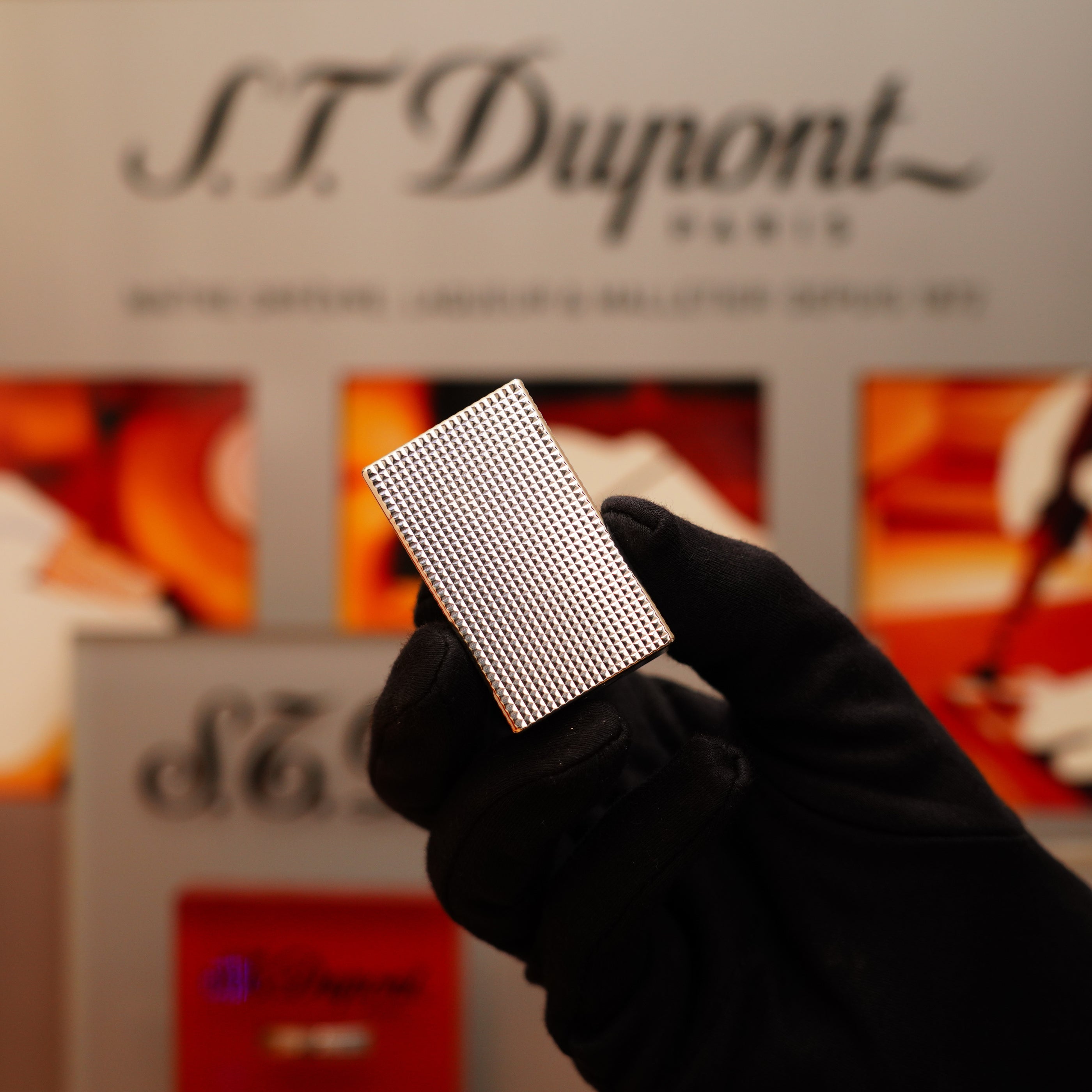 A gloved hand holds a rectangular, textured metallic object in front of a blurred S.T. Dupont Paris display. This is the Vintage 1996 St Dupont Ligne 1 Large Heavy Silver Finish Lighter with a Diamond Head Pattern by S.T. Dupont.