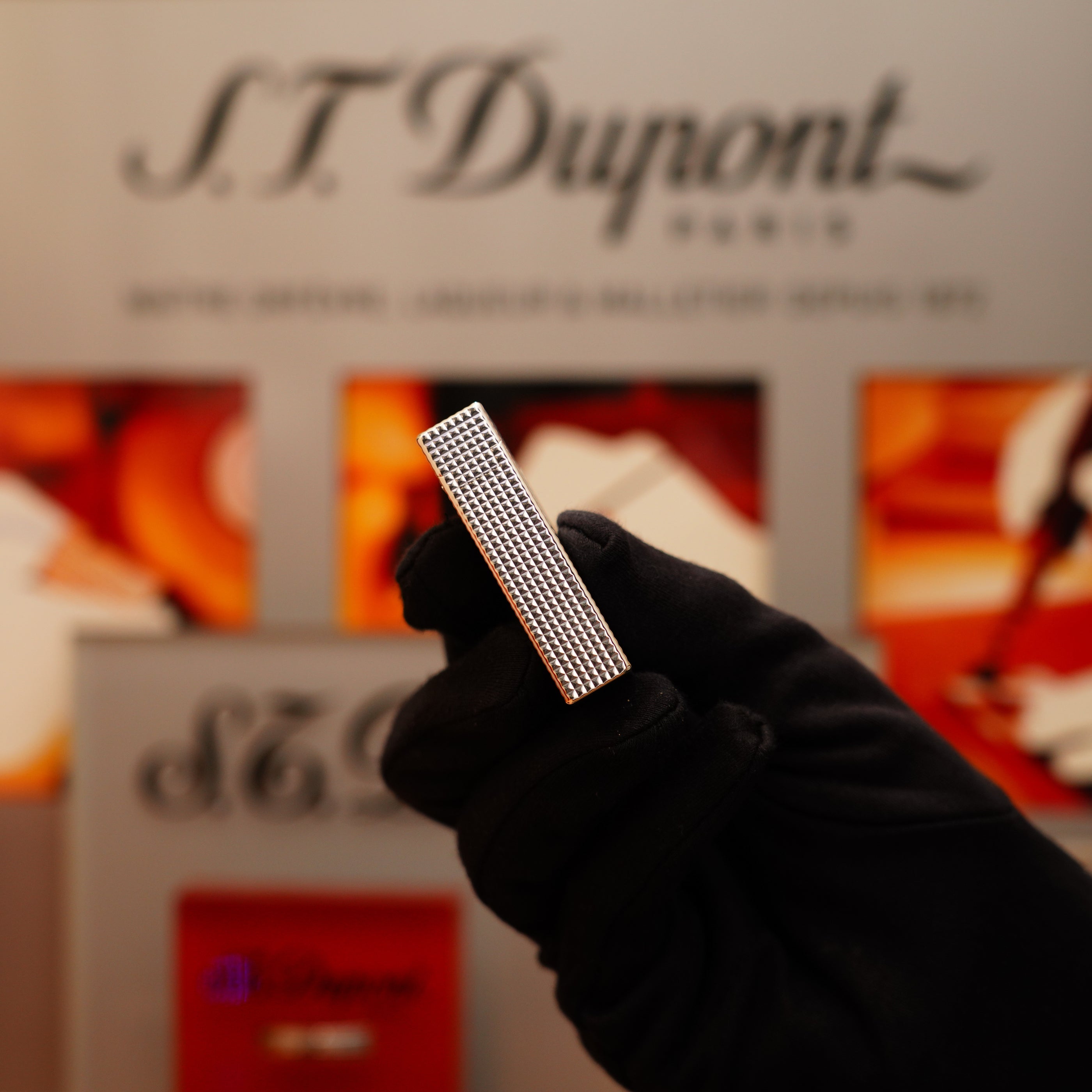 A gloved hand holds a rectangular, textured silver object, identified as the Vintage 1996 St Dupont Ligne 1 Diamond Head Pattern Large Heavy Silver Finish Lighter, with a blurred S.T. Dupont sign in the background.