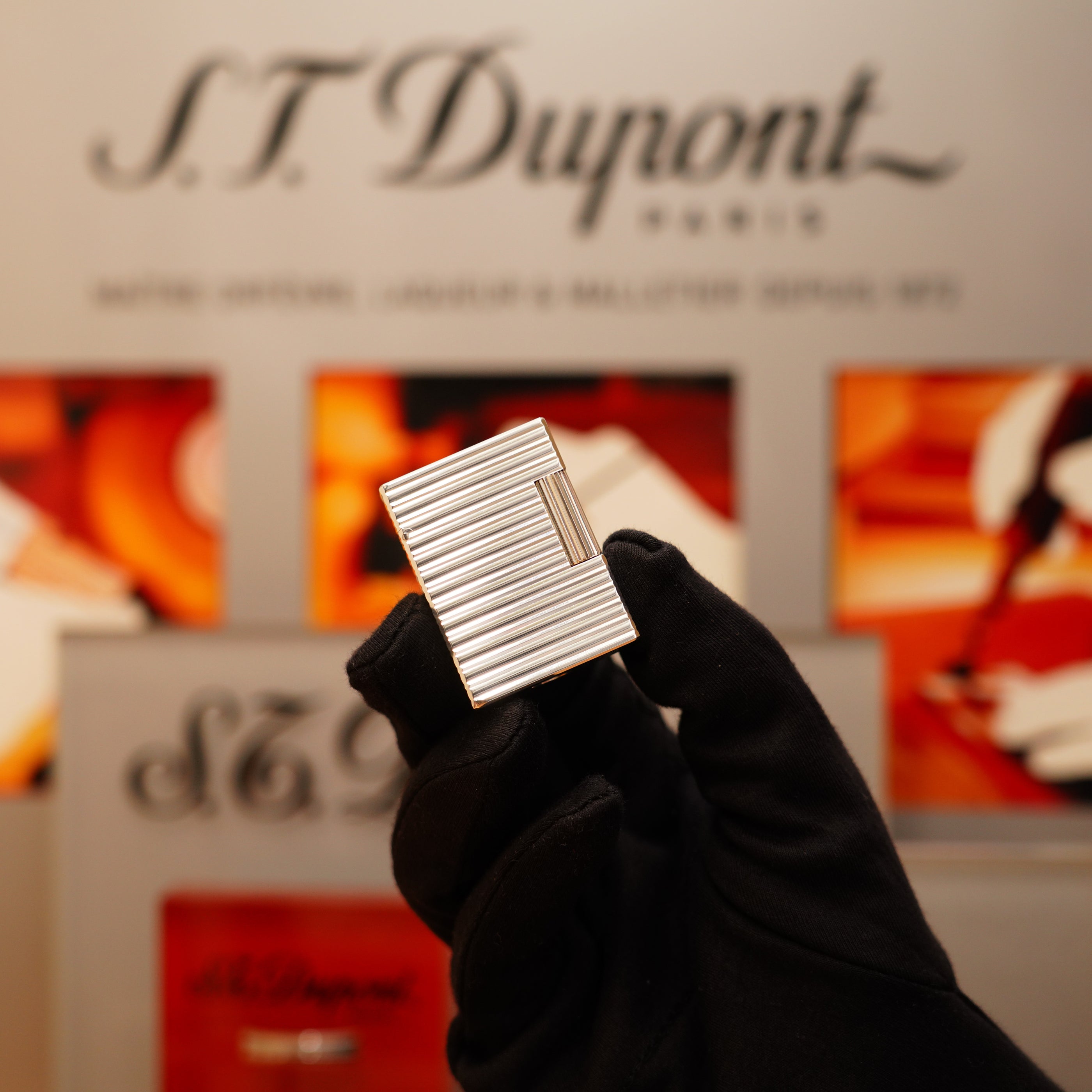 A gloved hand holds a Vintage 1996 S.T. Dupont Ligne 1 small Horizontal line Heavy Silver Finish Lighter against a blurred background with S.T. Dupont branding.