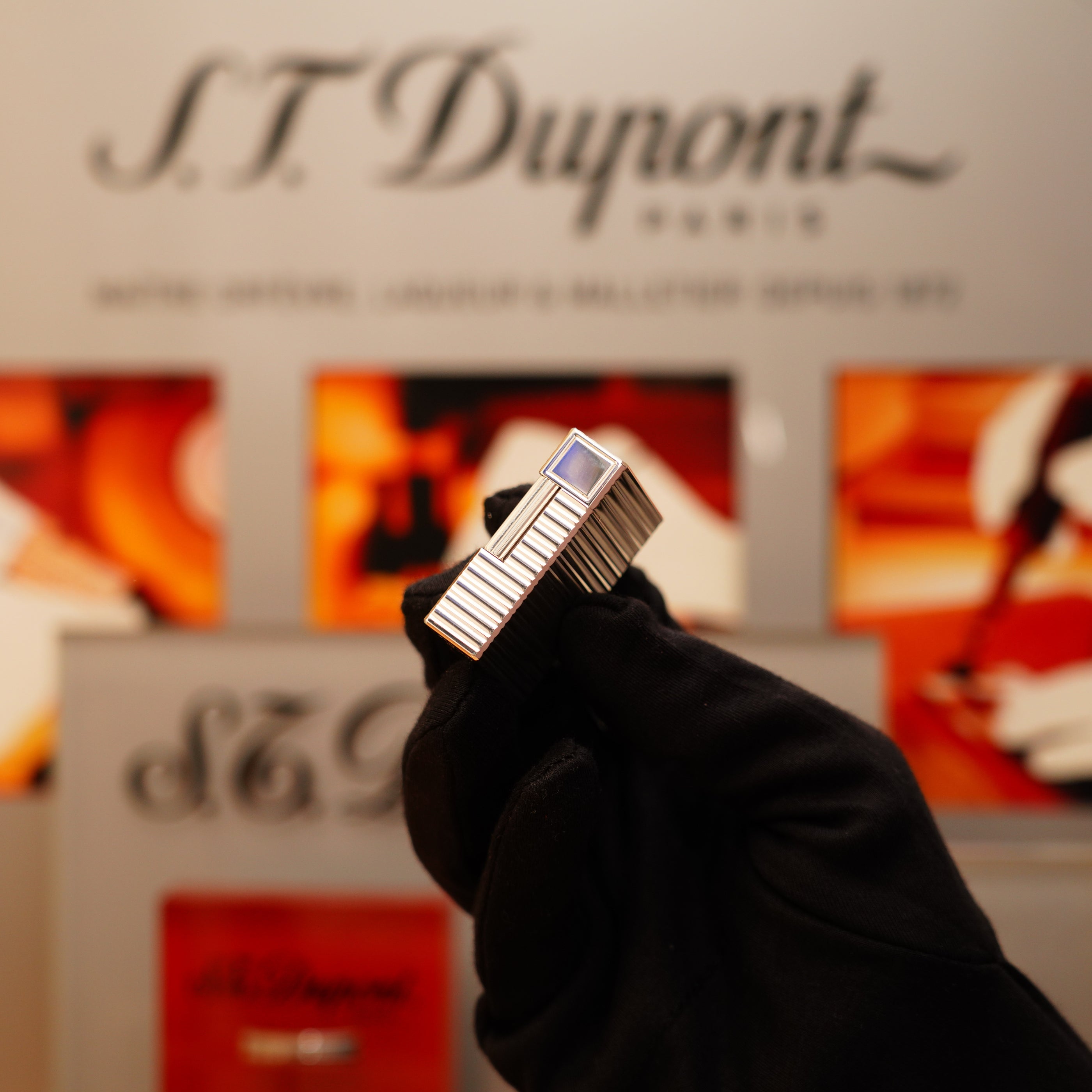 A gloved hand holds a Vintage 1996 S.T. Dupont Ligne 1 small Horizontal Line Heavy Silver Finish lighter against a branded background.