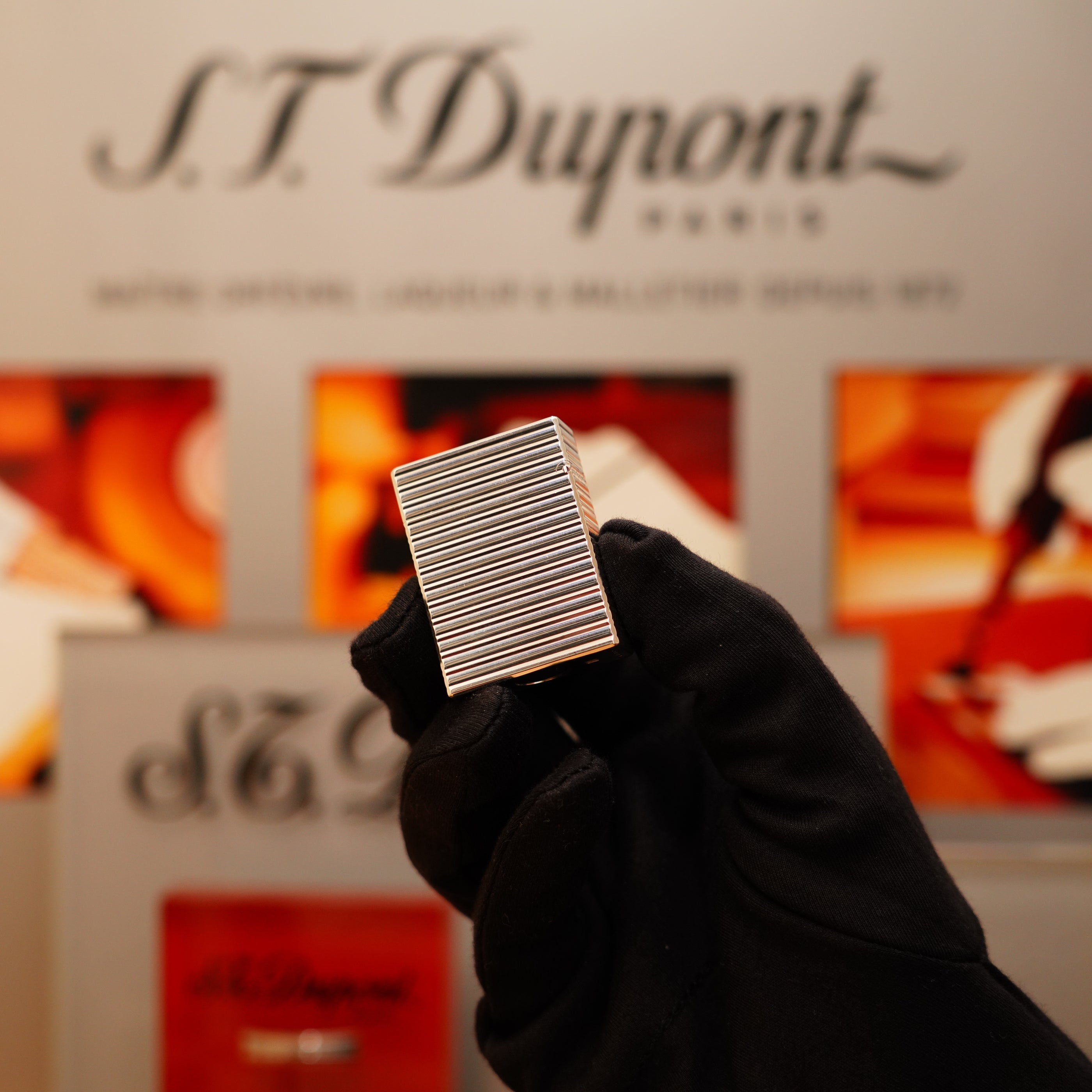 A gloved hand holds a Vintage 1996 S.T. Dupont Ligne 1 small lighter with a horizontal line heavy silver finish in front of a blurred S.T. Dupont display.