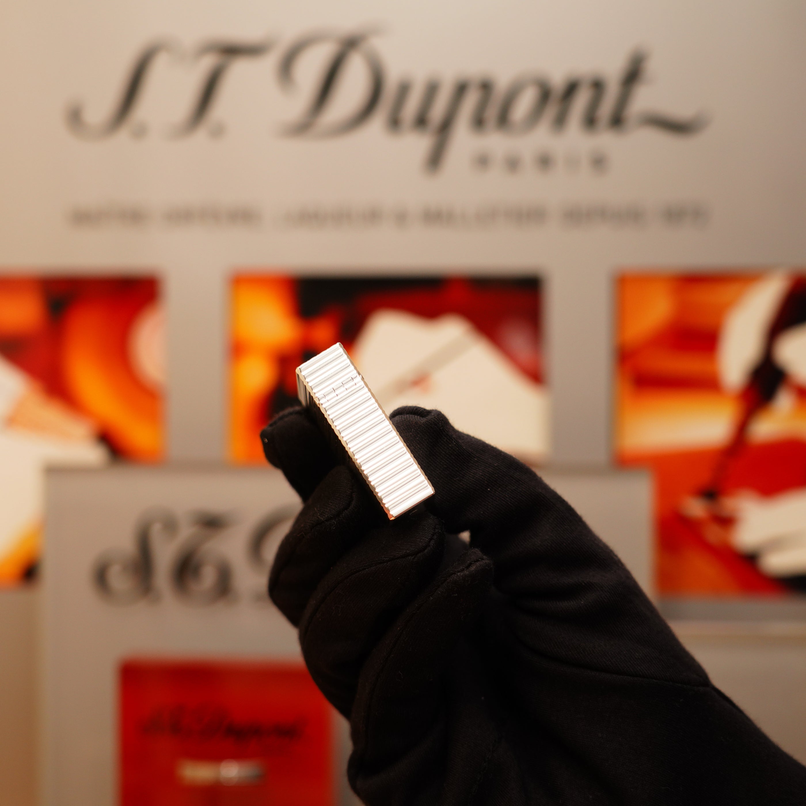 A gloved hand holds a Vintage 1996 S.T. Dupont Ligne 1 small lighter with a horizontal line heavy silver finish, set against a blurred S.T. Dupont Paris sign.