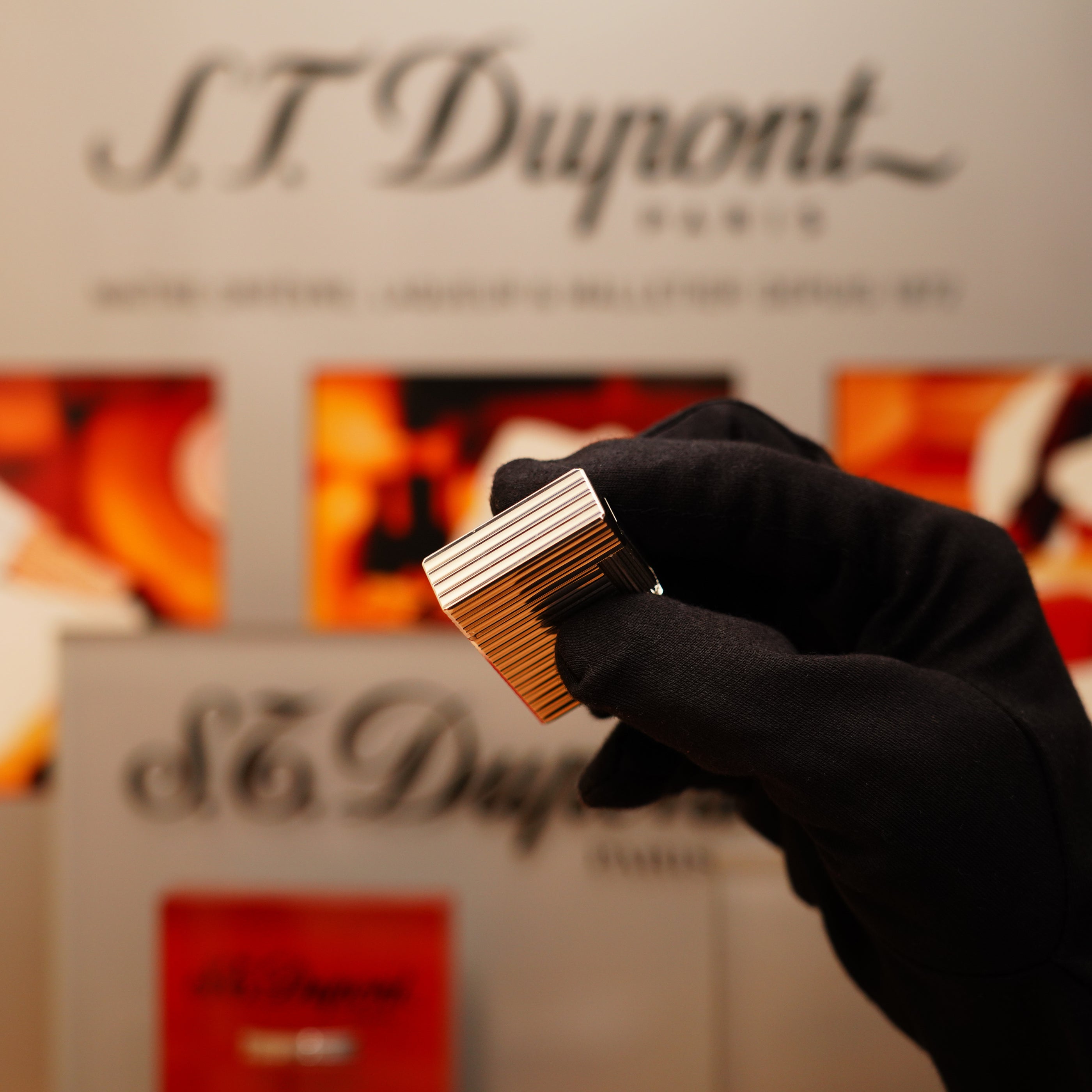 A gloved hand holds a Vintage 1996 S.T. Dupont Ligne 1 small Horizontal line Heavy Silver Finish Lighter in front of a blurred S.T. Dupont signage.