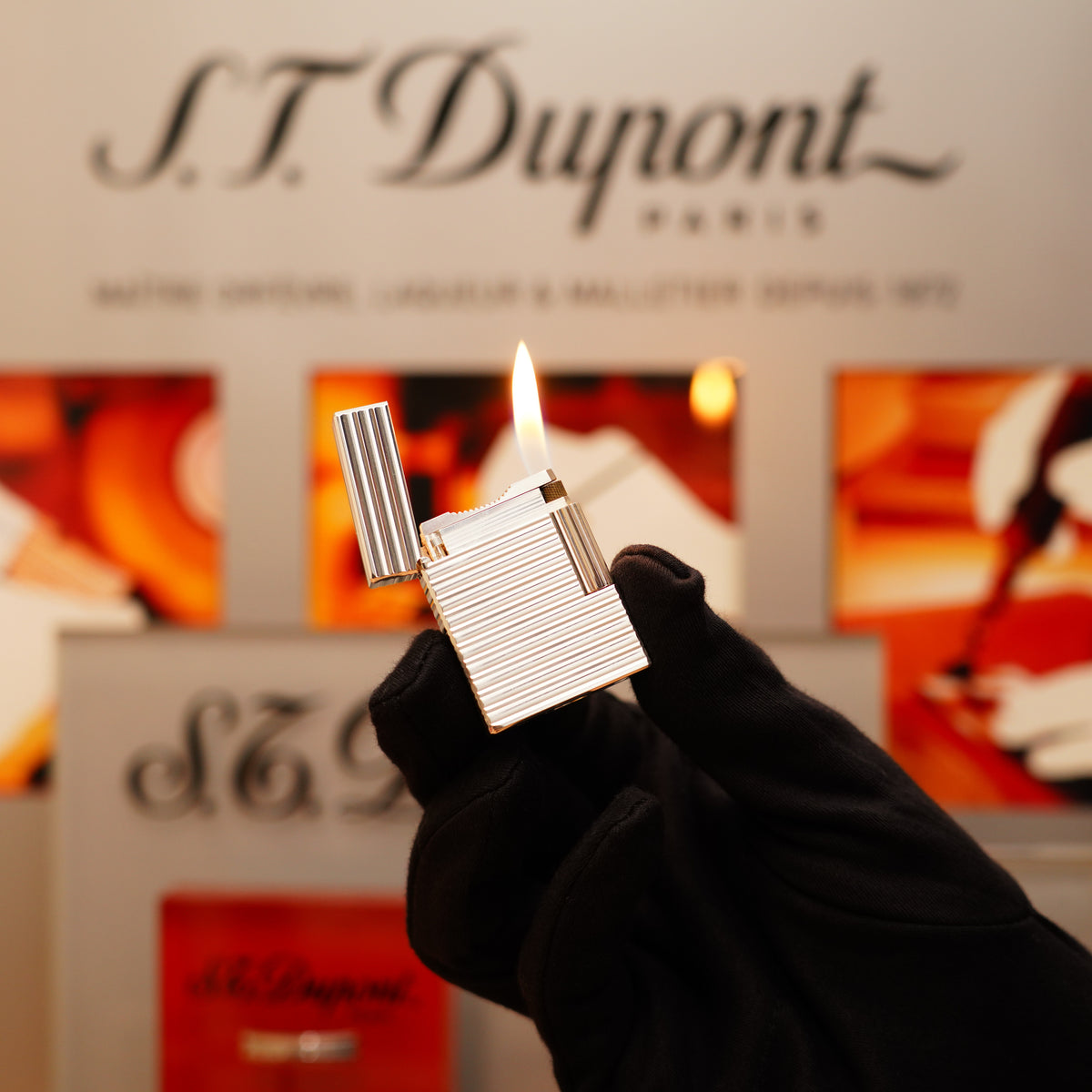 A gloved hand holds a lit Vintage 1996 S.T. Dupont Ligne 1 Small Horizontal Line Heavy Silver Finish Lighter with the S.T. Dupont logo visible in the background.