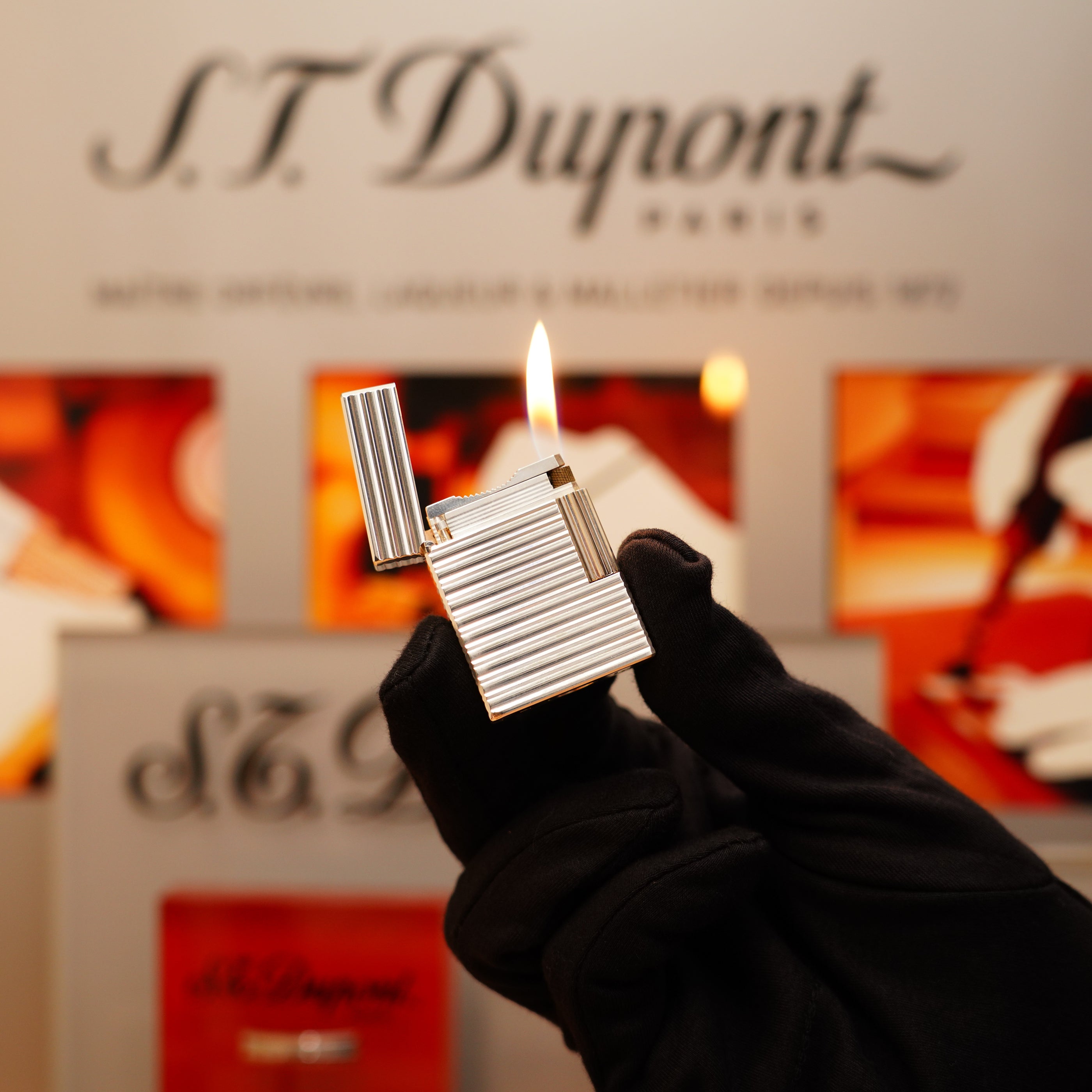 A hand in a black glove holds a lit Vintage 1996 S.T. Dupont Ligne 1 small lighter with a corrugated heavy silver finish, set against the background featuring the S.T. Dupont logo and blurred images.
