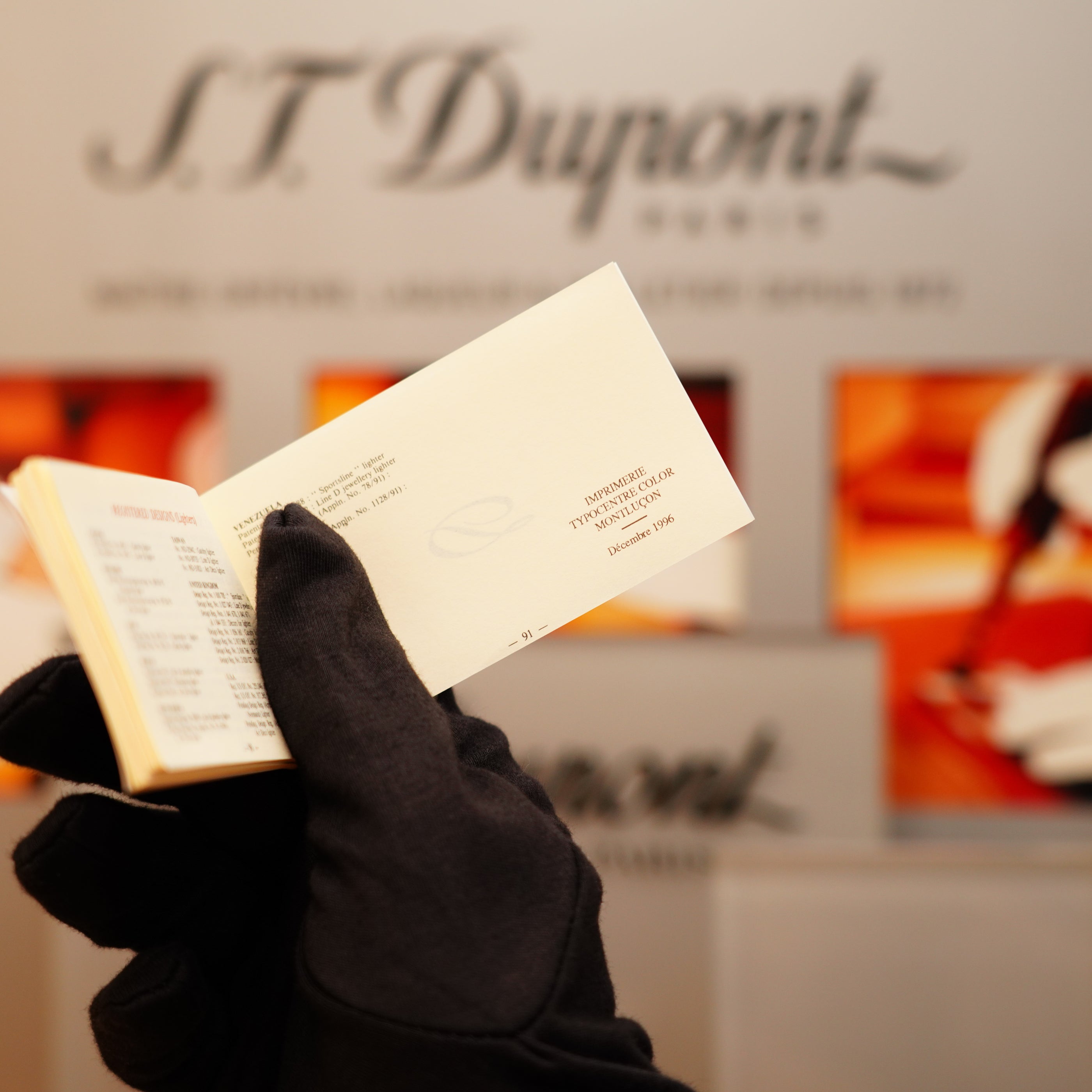A hand in a black glove holds a compact item against a blurred background, showcasing the brand name "S.T. Dupont.