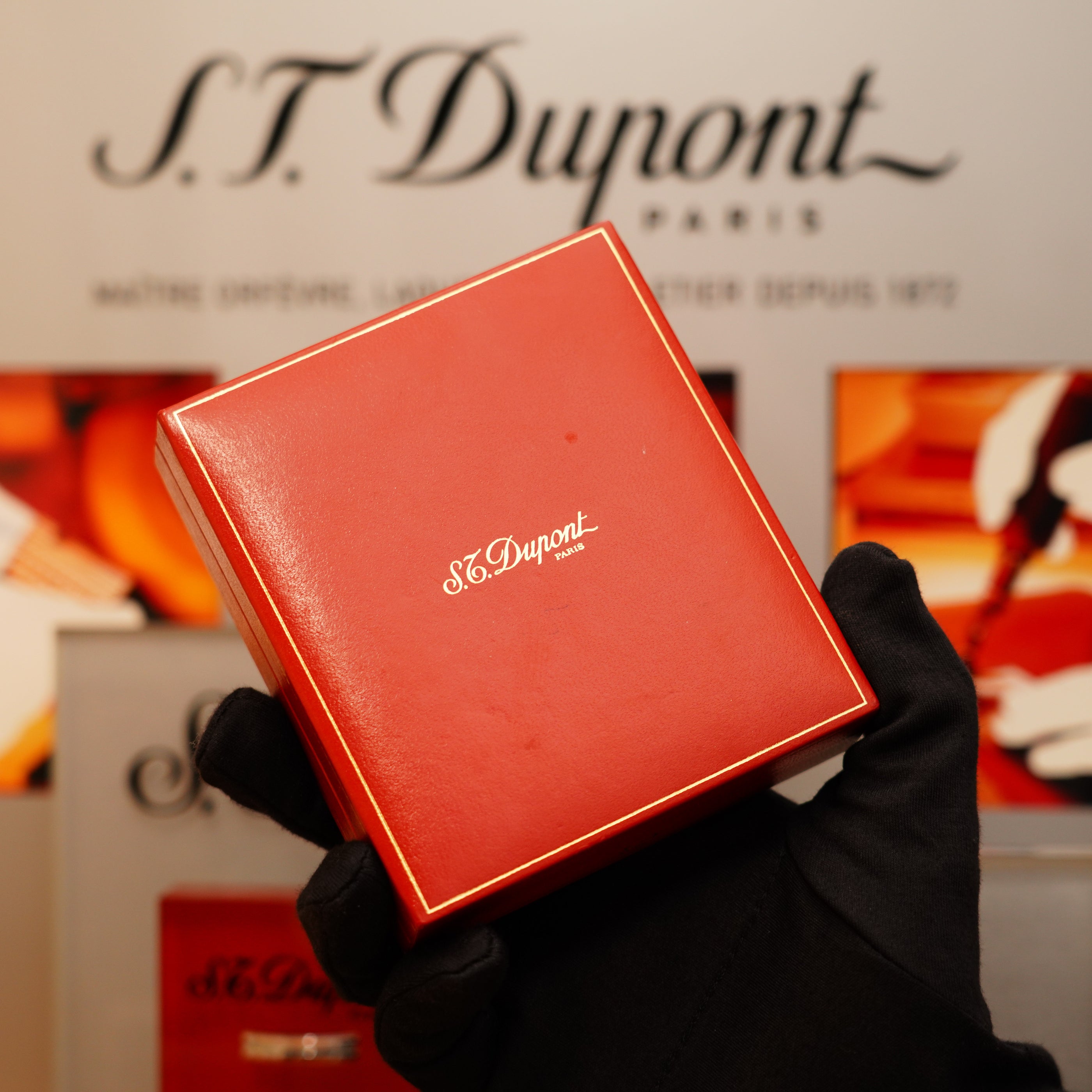 A gloved hand holds a red box with "S.T. Dupont" printed on it, in front of a blurred background featuring the brand name and showcasing the Vintage 1996 St Dupont Ligne 1 small Horizontal line Heavy Silver Finish Lighter.