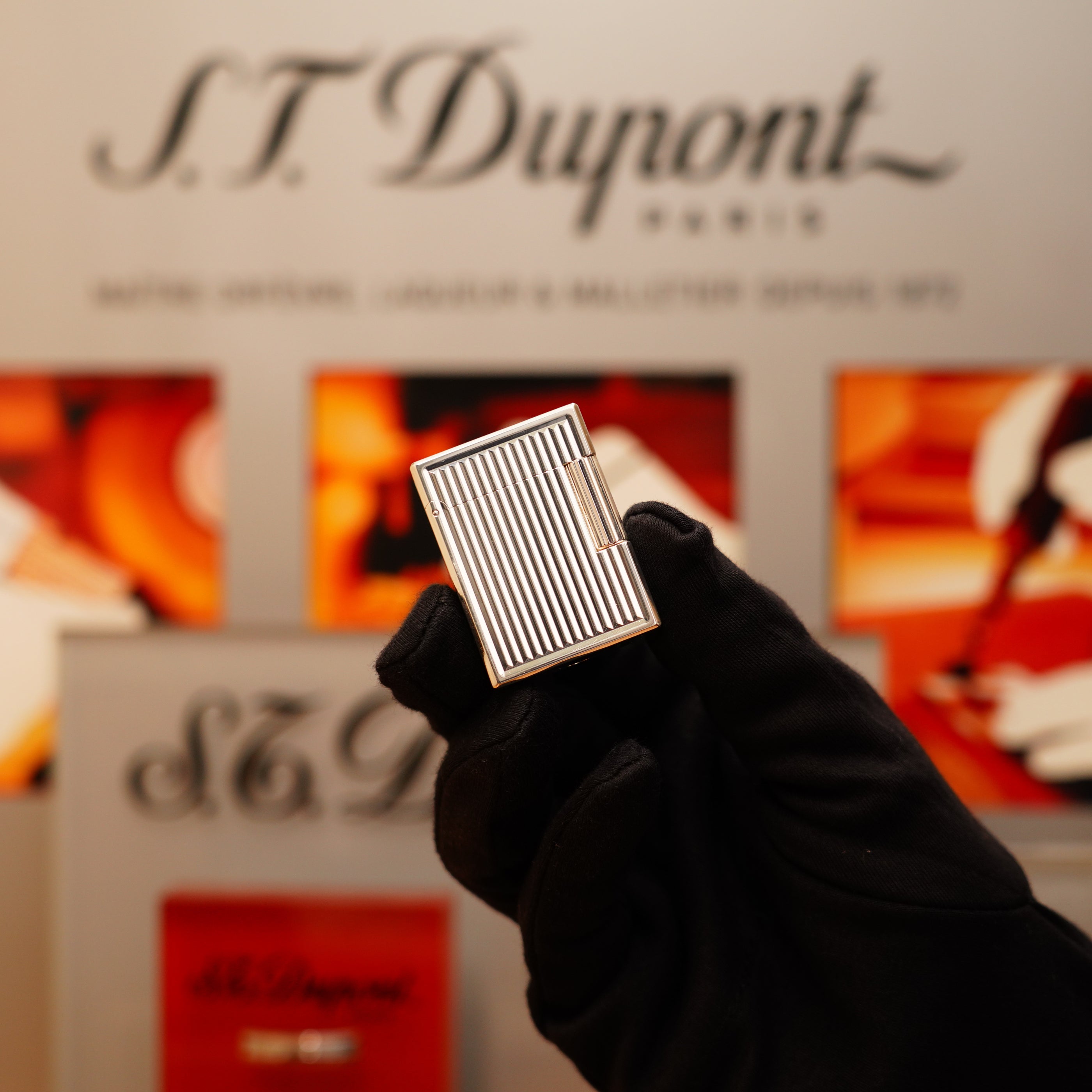 A gloved hand holds an S.T. Dupont lighter, identified as the 1996 Vintage Ligne 1 with a heavy silver finish and vertical line pattern, against a blurred background featuring logos and artwork.