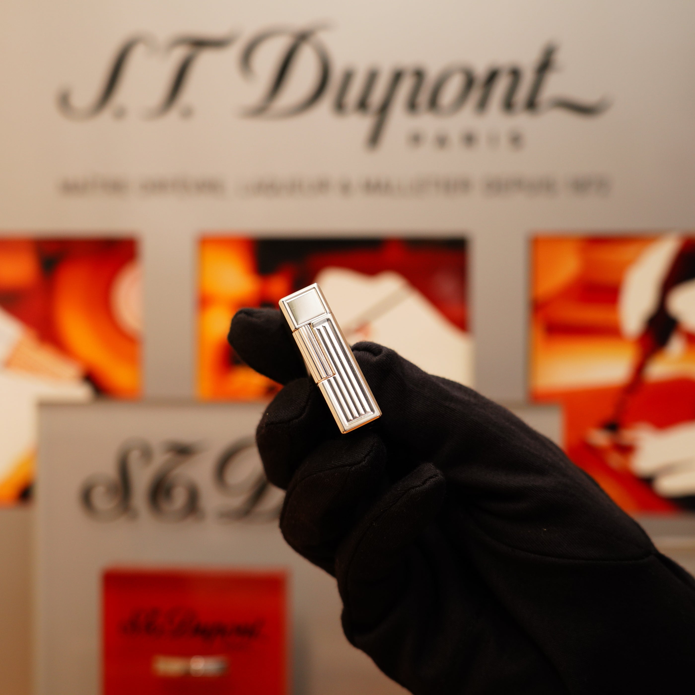 A gloved hand holds a Vintage 1996 S.T. Dupont Ligne 1 lighter with a rare heavy silver finish and vertical line pattern, in front of a display featuring the brand's logo and images.