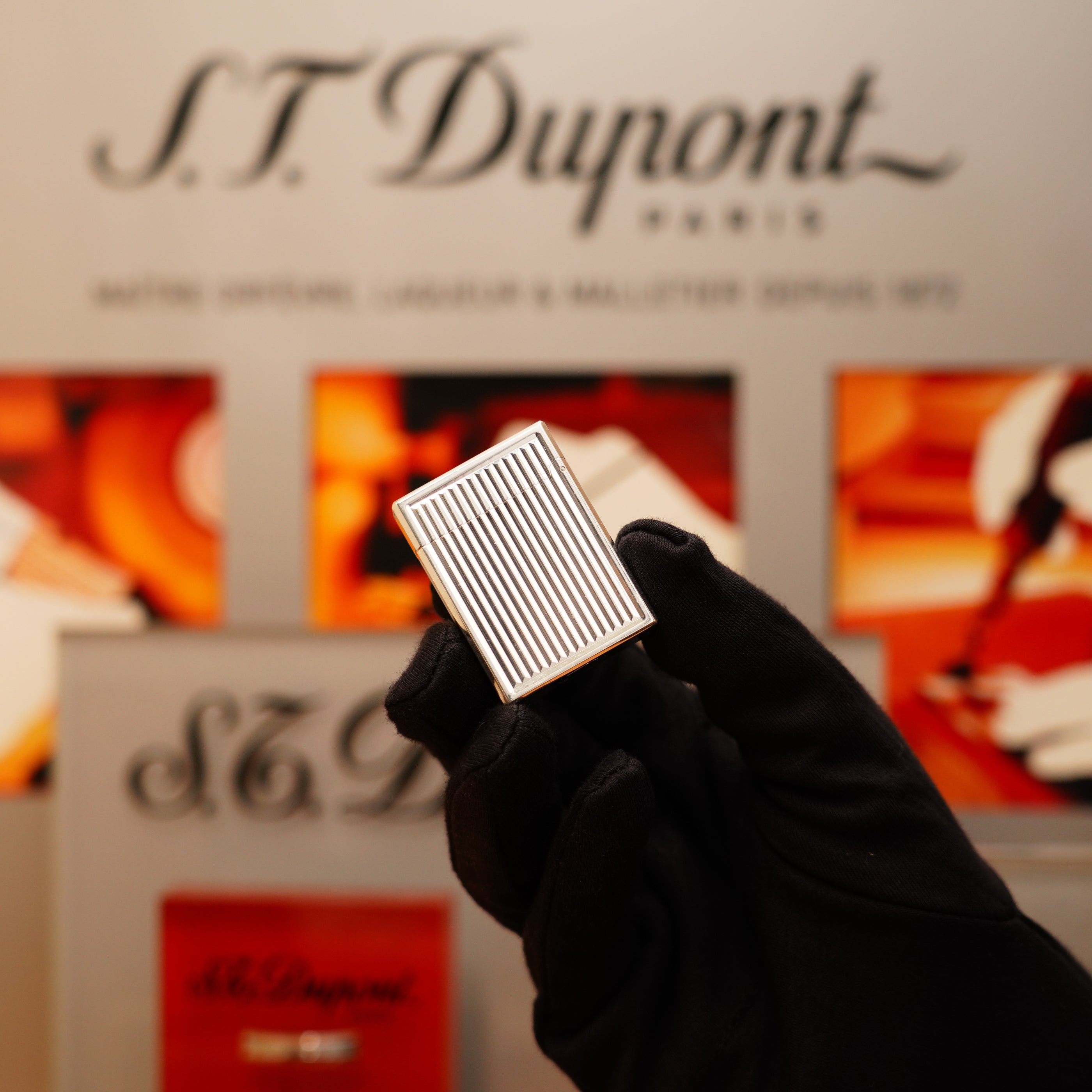 A gloved hand holds a Vintage 1996 S.T. Dupont Ligne 1 lighter, featuring a rare heavy silver finish and vertical line pattern, in front of a blurred S.T. Dupont Paris sign.