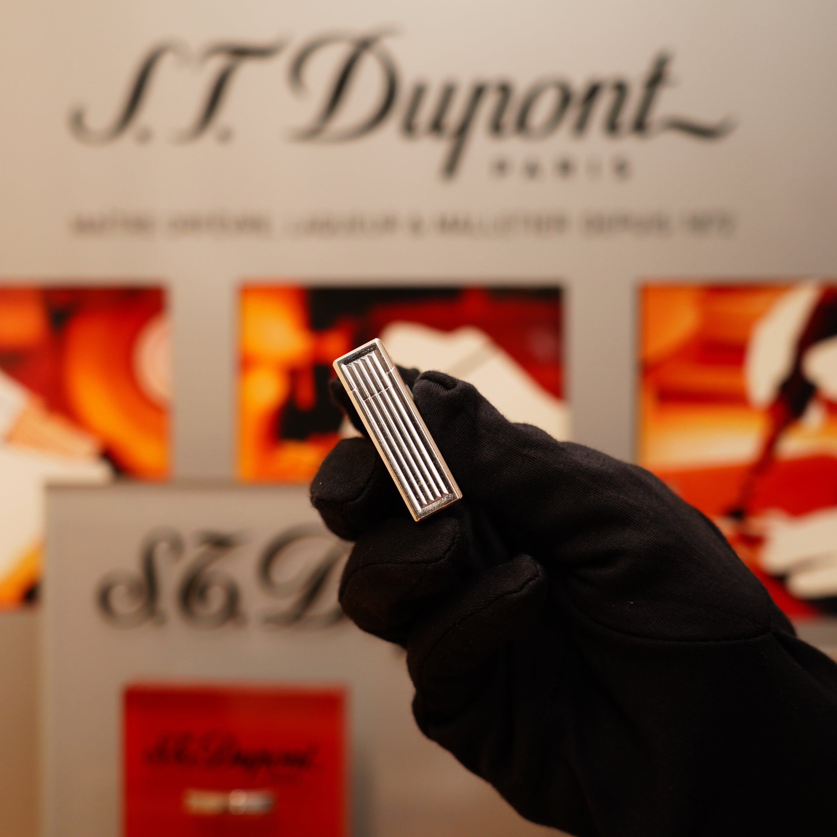 A person wearing black gloves is holding a Vintage 1996 S.T. Dupont Ligne 1 lighter with a rare, heavy silver finish and vertical line pattern in front of an S.T. Dupont display background.