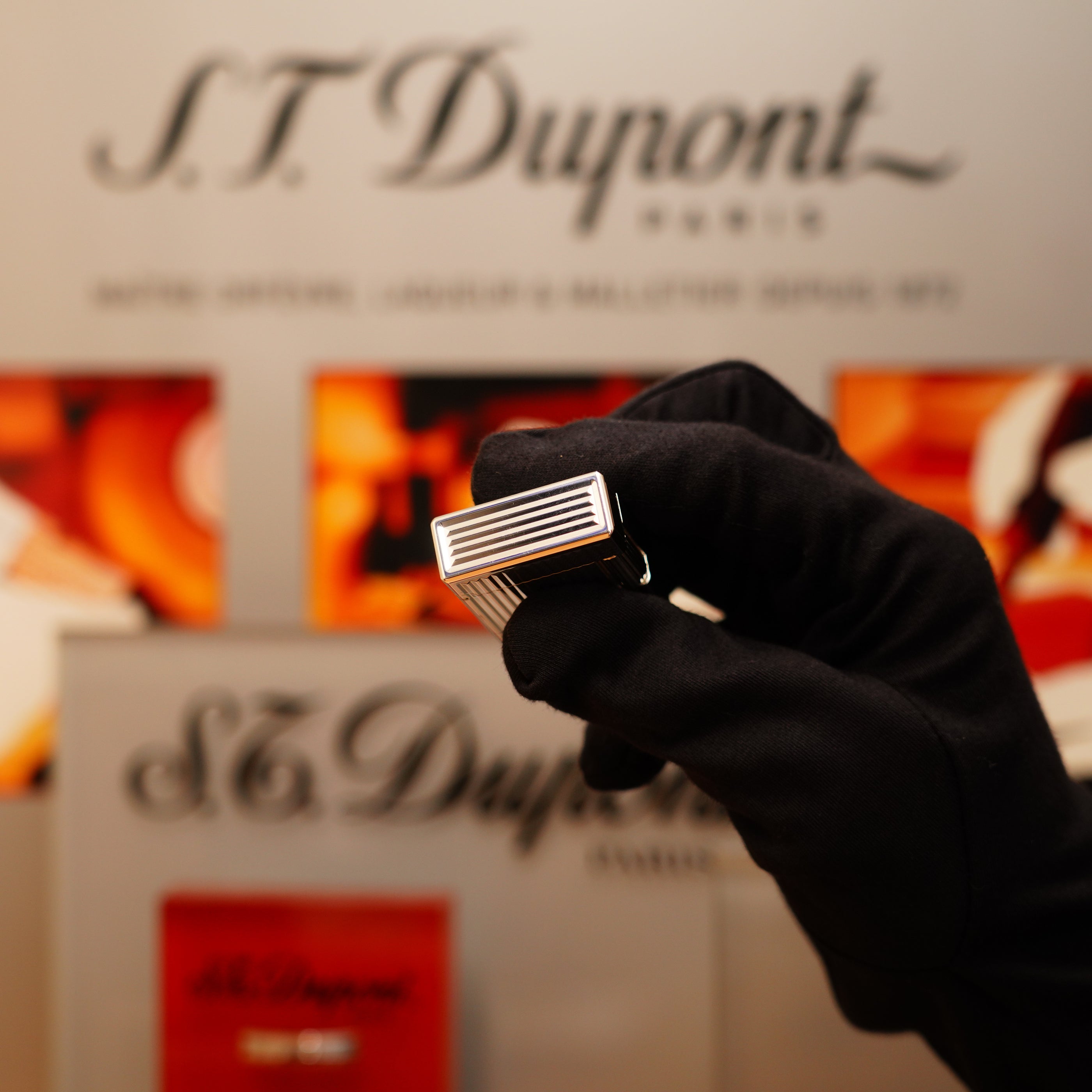 A gloved hand holds a Vintage 1996 S.T. Dupont Ligne 1 Rare Heavy Silver Finish Vertical Line Pattern Lighter in front of a blurred S.T. Dupont logo backdrop.
