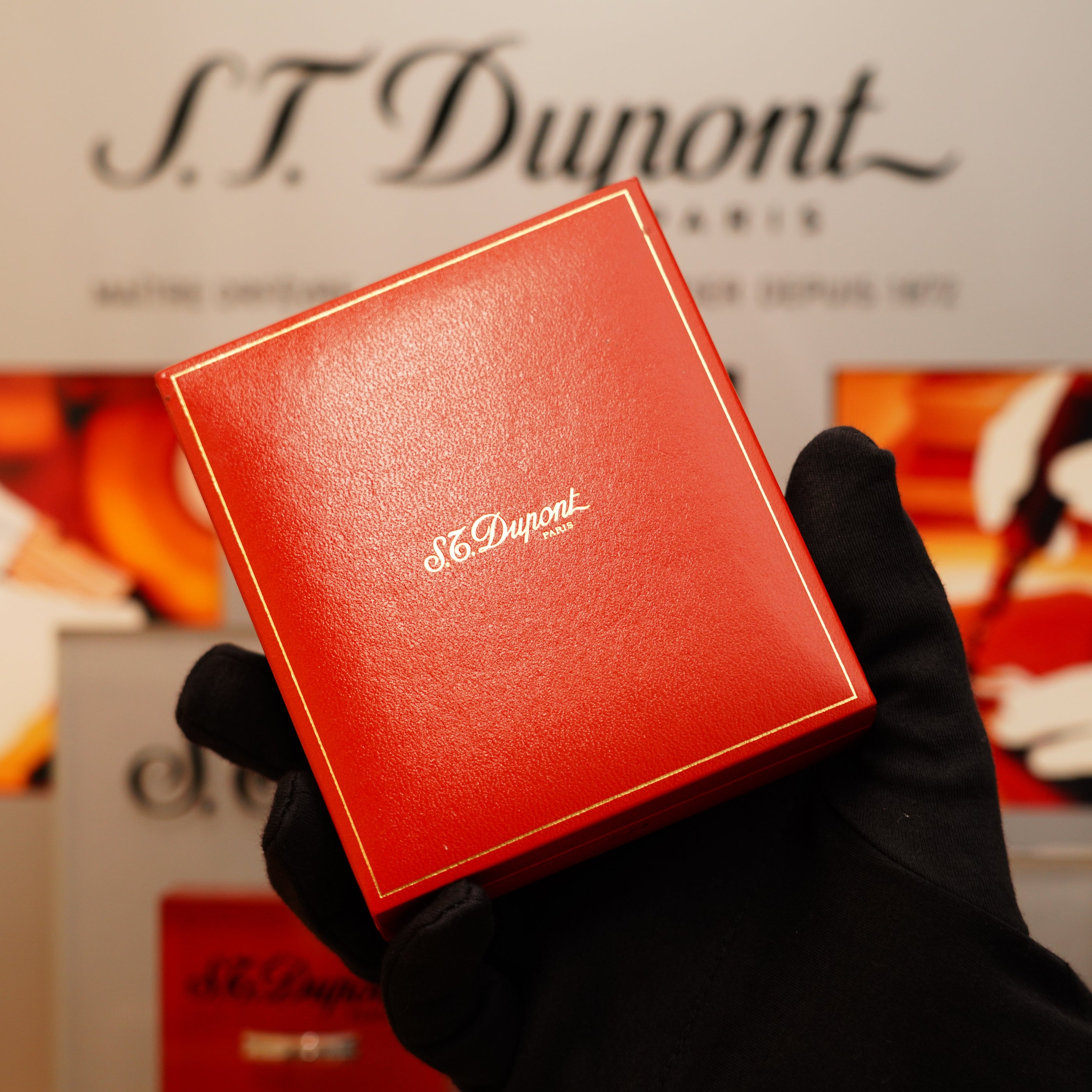 A gloved hand displays a red S.T. Dupont box with gold trim, set against a blurred background featuring the S.T. Dupont logo, highlighting the Vintage 1996 St Dupont Ligne 1 Rare Heavy Silver Finish Vertical Line Pattern Lighter.