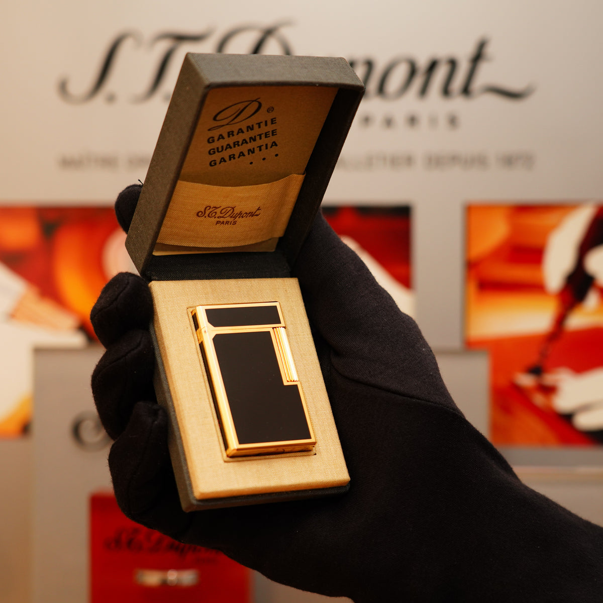 A black-gloved hand presents an open box with a Vintage 1973 S.T. Dupont Japanese Lac Signed 24k Heavy Gold Large Ligne 1 Natural Full Black Lacquer Iconic Model Lighter inside, set against a blurred background.