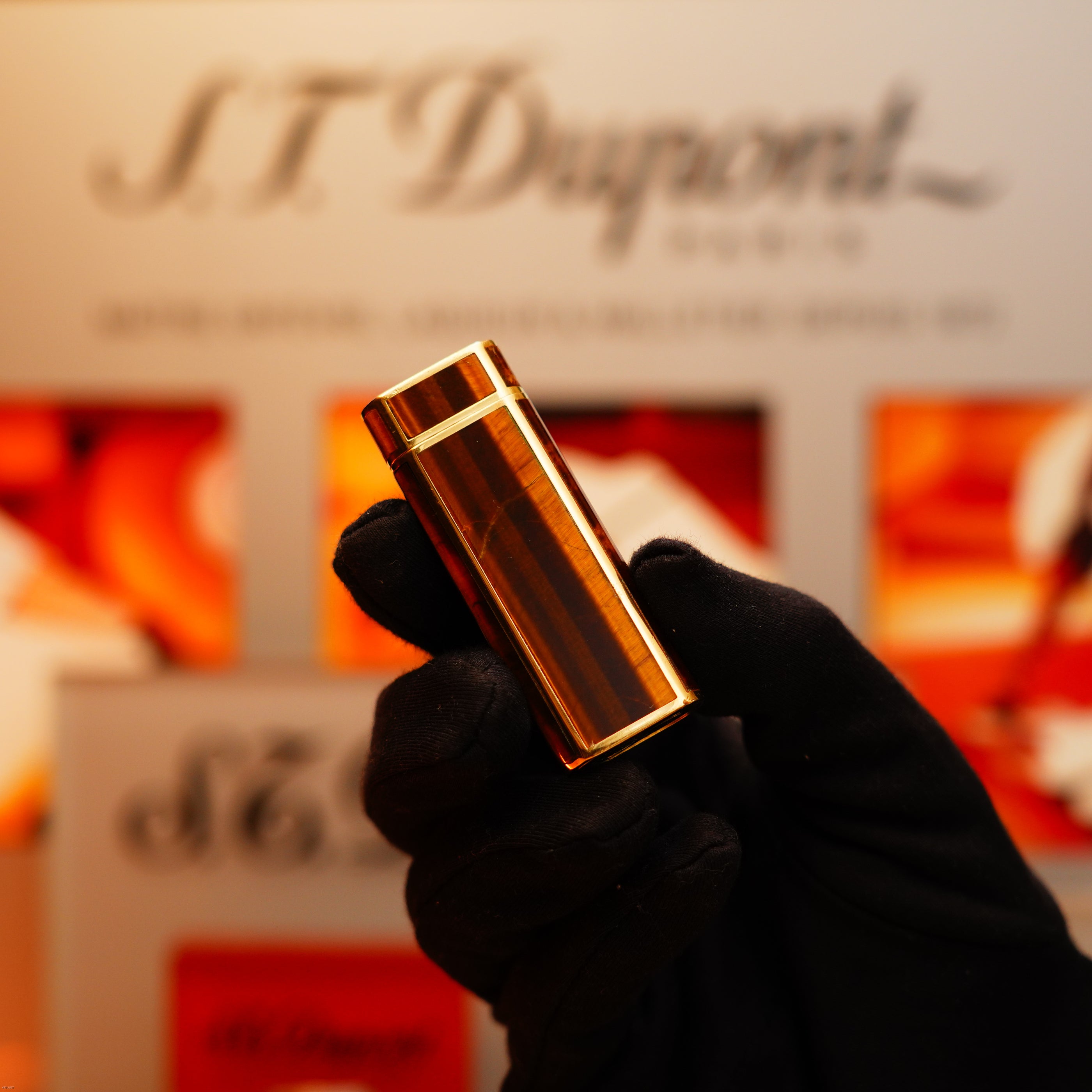 A gloved hand holds a Vintage 1970 Cartier Extremely Rare Tiger's Eye Solid 18k Lighter with 750 hallmark from KirbyAllison.com in front of a blurred branded display.