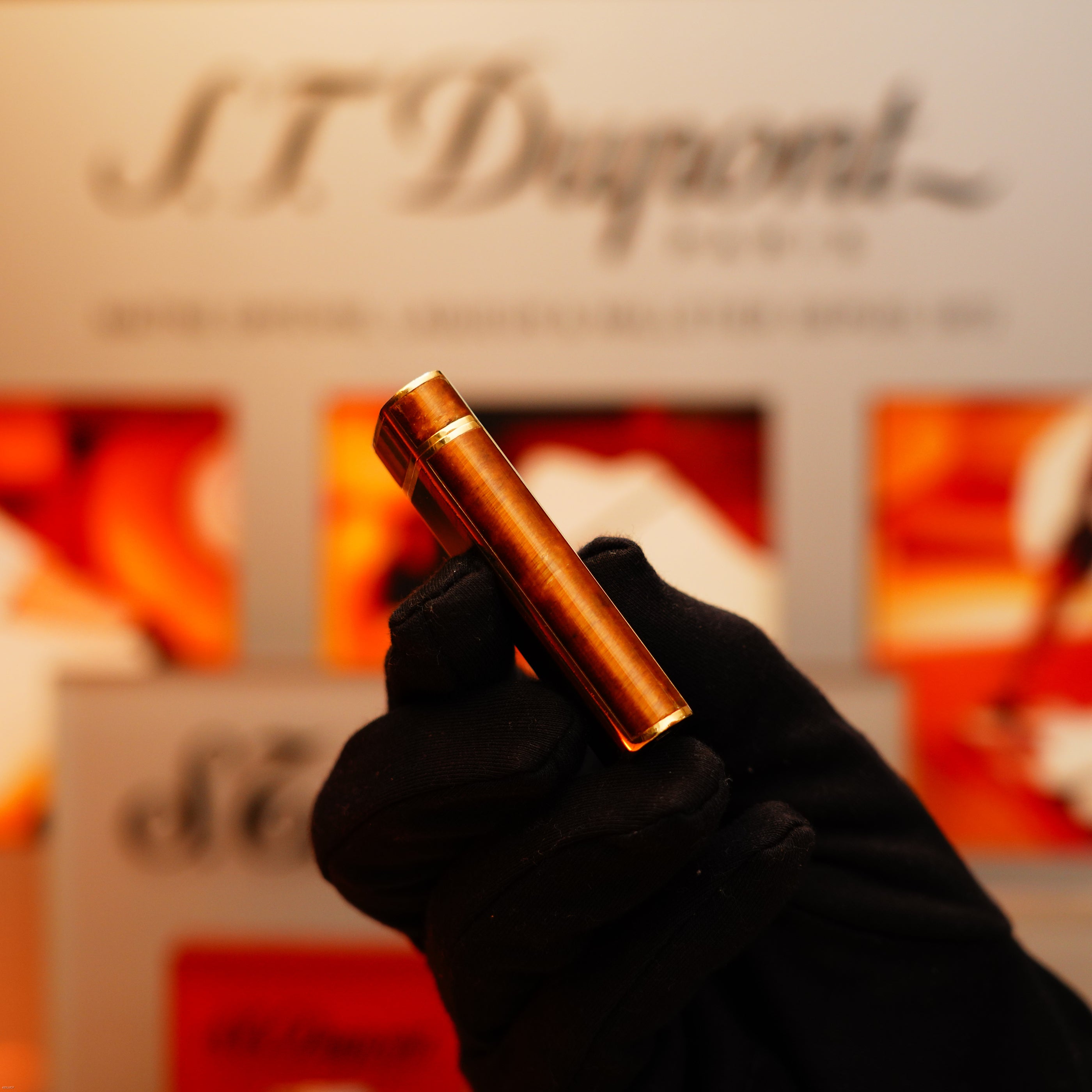 A gloved hand holds a Vintage 1970 Cartier Tiger's Eye Solid 18k Lighter (750 hallmark) from KirbyAllison.com before a blurred display.