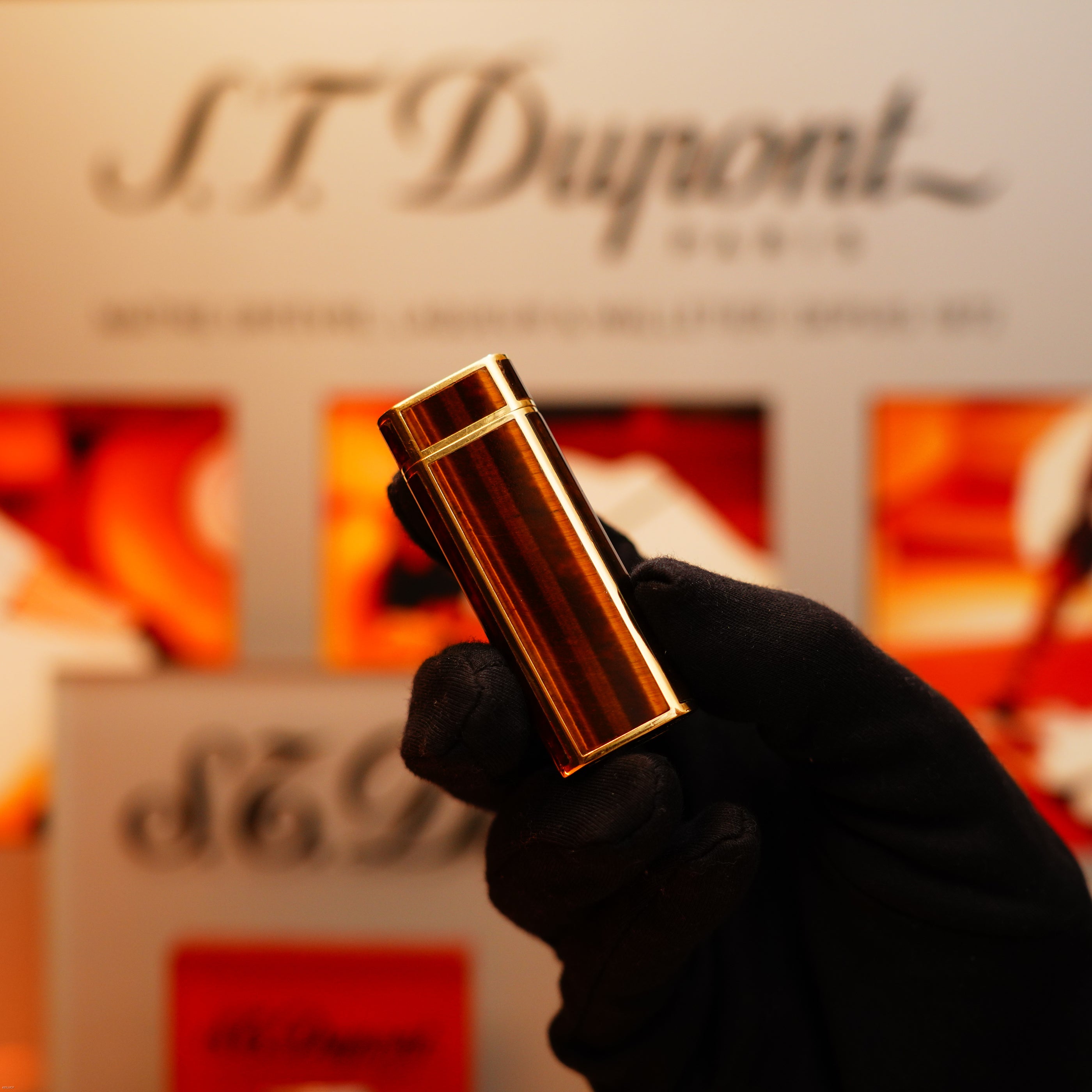 A gloved hand holds a Vintage 1970 Cartier Extremely Rare Tiger's Eye Solid 18k Heavy Body Lighter with a 750 hallmark from KirbyAllison.com, with a blurred branded display in the background.