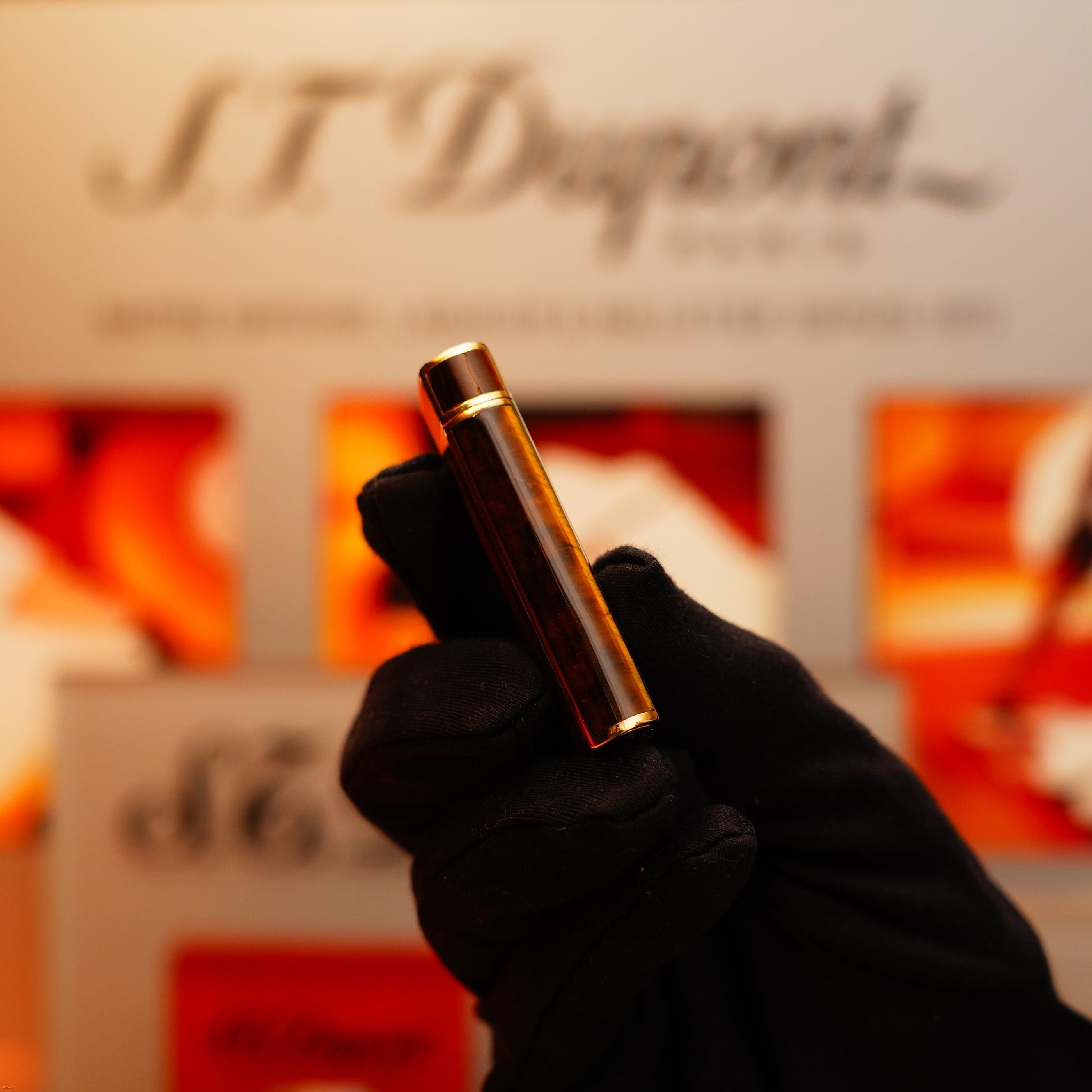 A gloved hand holds a Vintage 1970 Cartier Tiger's Eye Solid 18k Lighter with 750 hallmark from KirbyAllison.com, upright in front of a blurred display with orange accents.