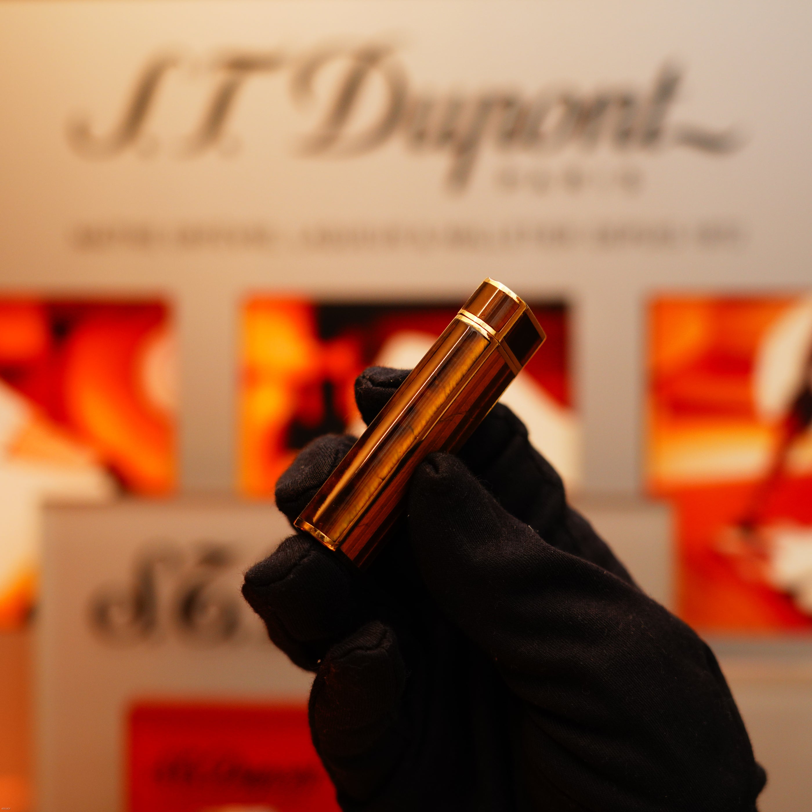 A gloved hand holds a Vintage 1970 Cartier Tiger's Eye Solid 18k Lighter with a 750 hallmark, by KirbyAllison.com, in front of a blurred display background.