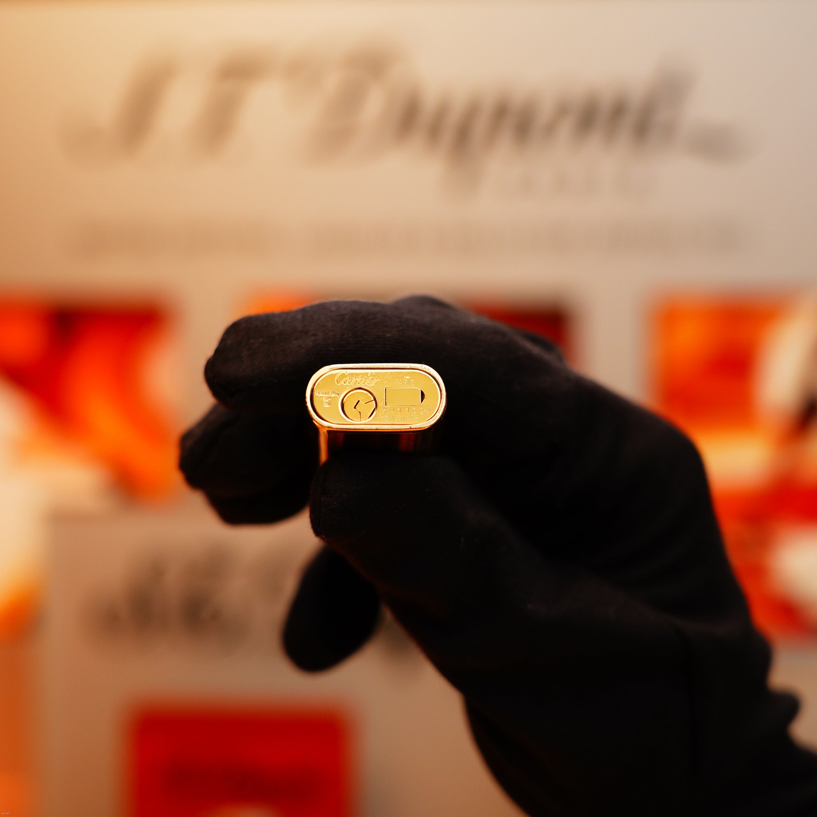 A person wearing a black glove holds a Vintage 1970 Cartier Extremely Rare Tiger's Eye Solid 18k Lighter with 750 hallmark, sold by KirbyAllison.com, with the branding visible in the blurred background.