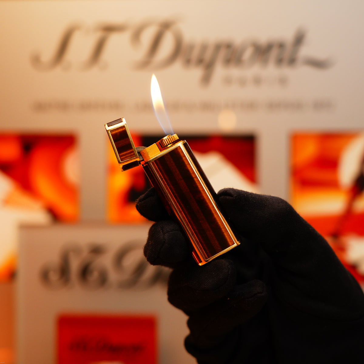 A gloved hand holds a lit Vintage 1970 Cartier Tiger's Eye Solid 18k lighter with 750 hallmark from KirbyAllison.com against a blurred background.