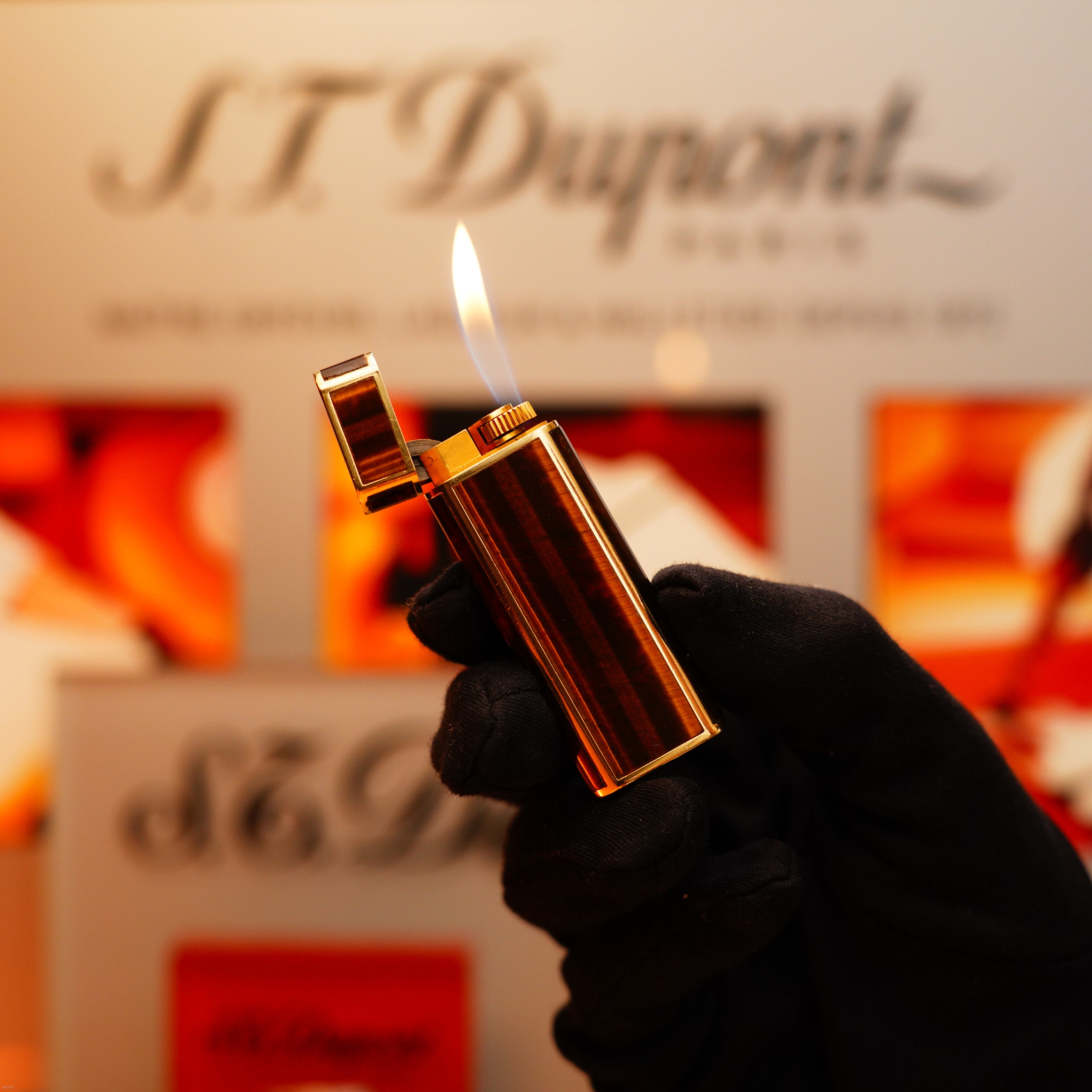 A gloved hand holds a lit Vintage 1970 Cartier Tiger's Eye Solid 18k lighter with 750 hallmark from KirbyAllison.com against a blurred background.