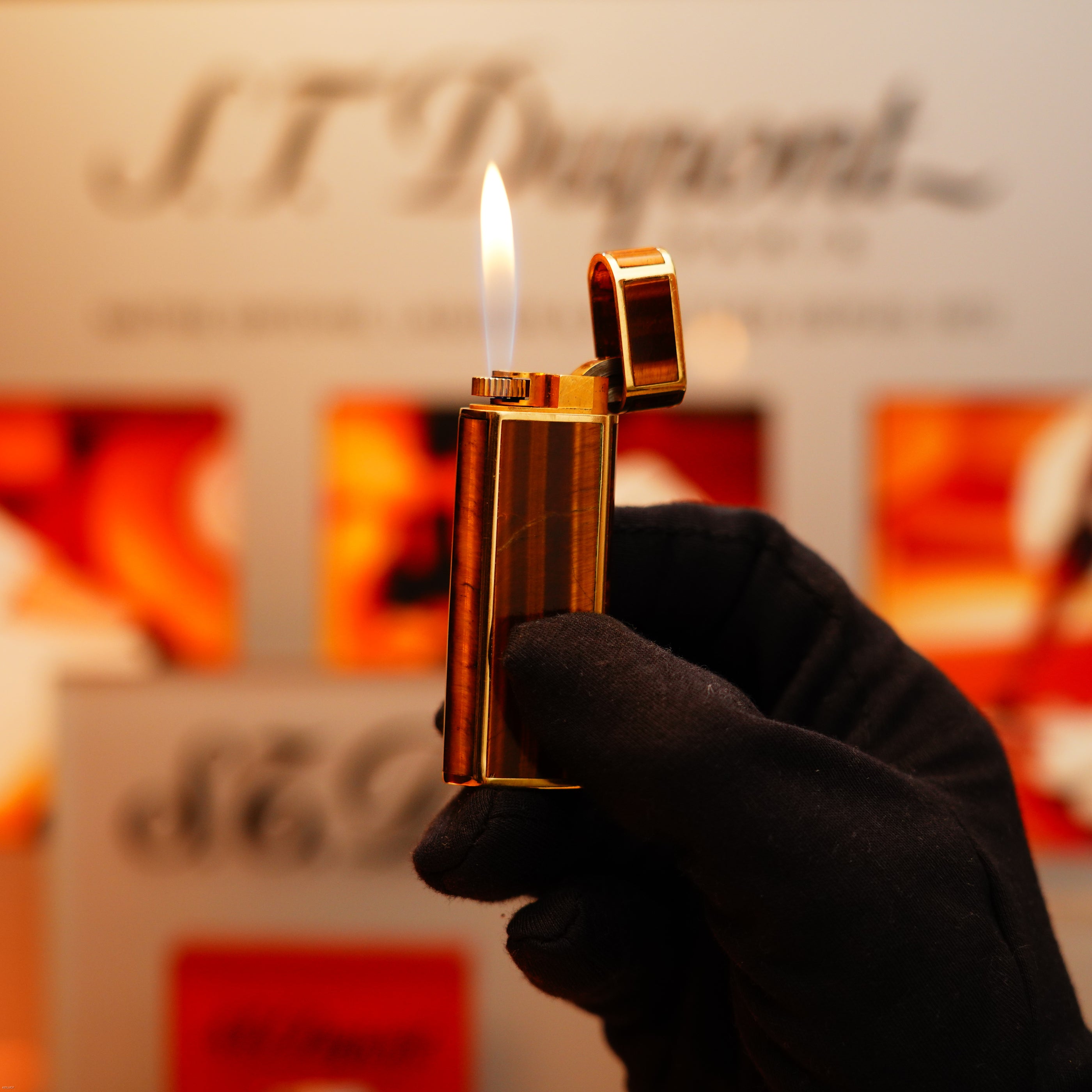 A gloved hand holds a lit Vintage 1970 Cartier Tiger's Eye Solid 18k Lighter—an extremely rare piece with a 750 hallmark—from KirbyAllison.com, with its flame glowing against a blurred branded background.