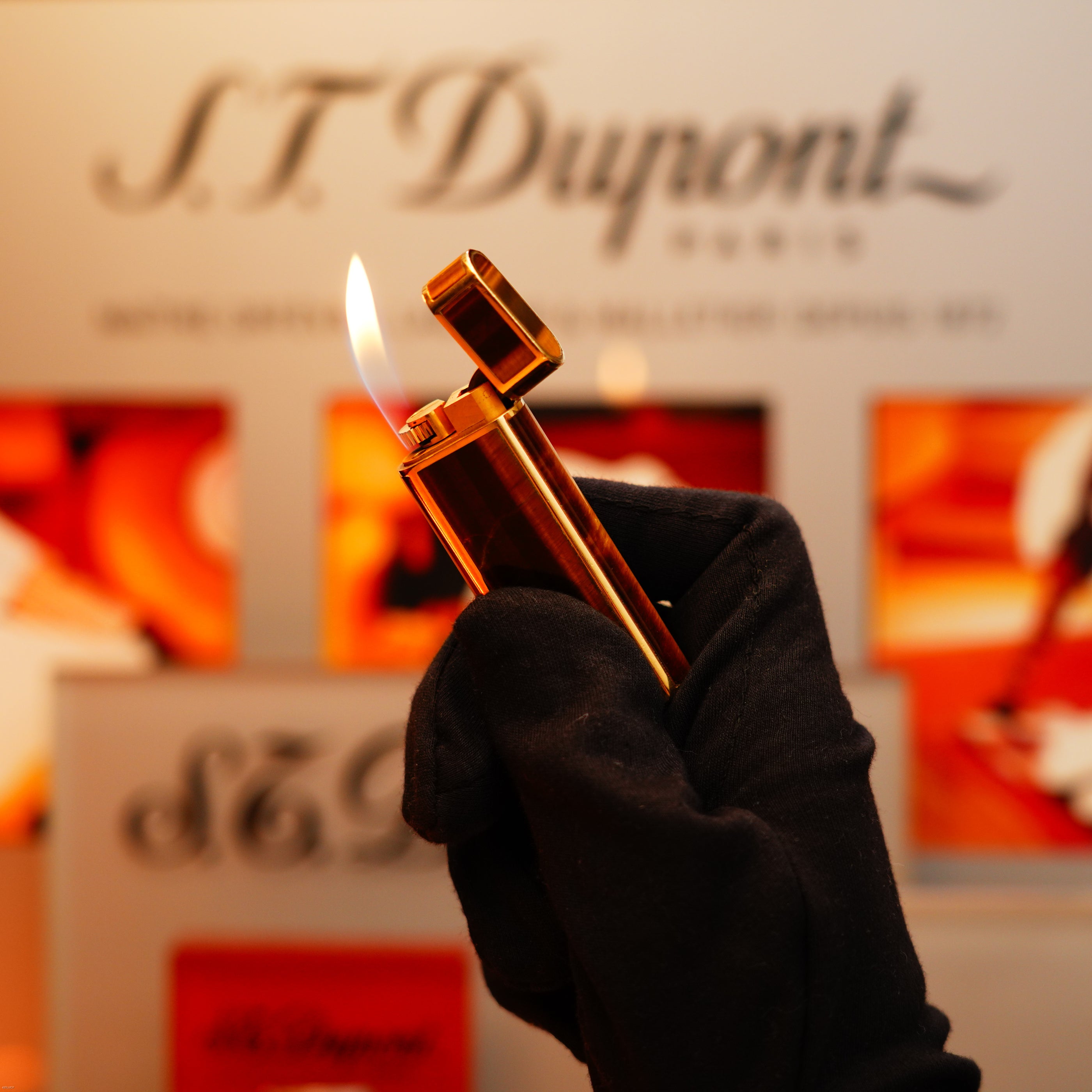A gloved hand holds a lit Vintage 1970 Cartier Tiger’s Eye Solid 18k Lighter with 750 hallmark from KirbyAllison.com, a blurred branded display visible in the background.