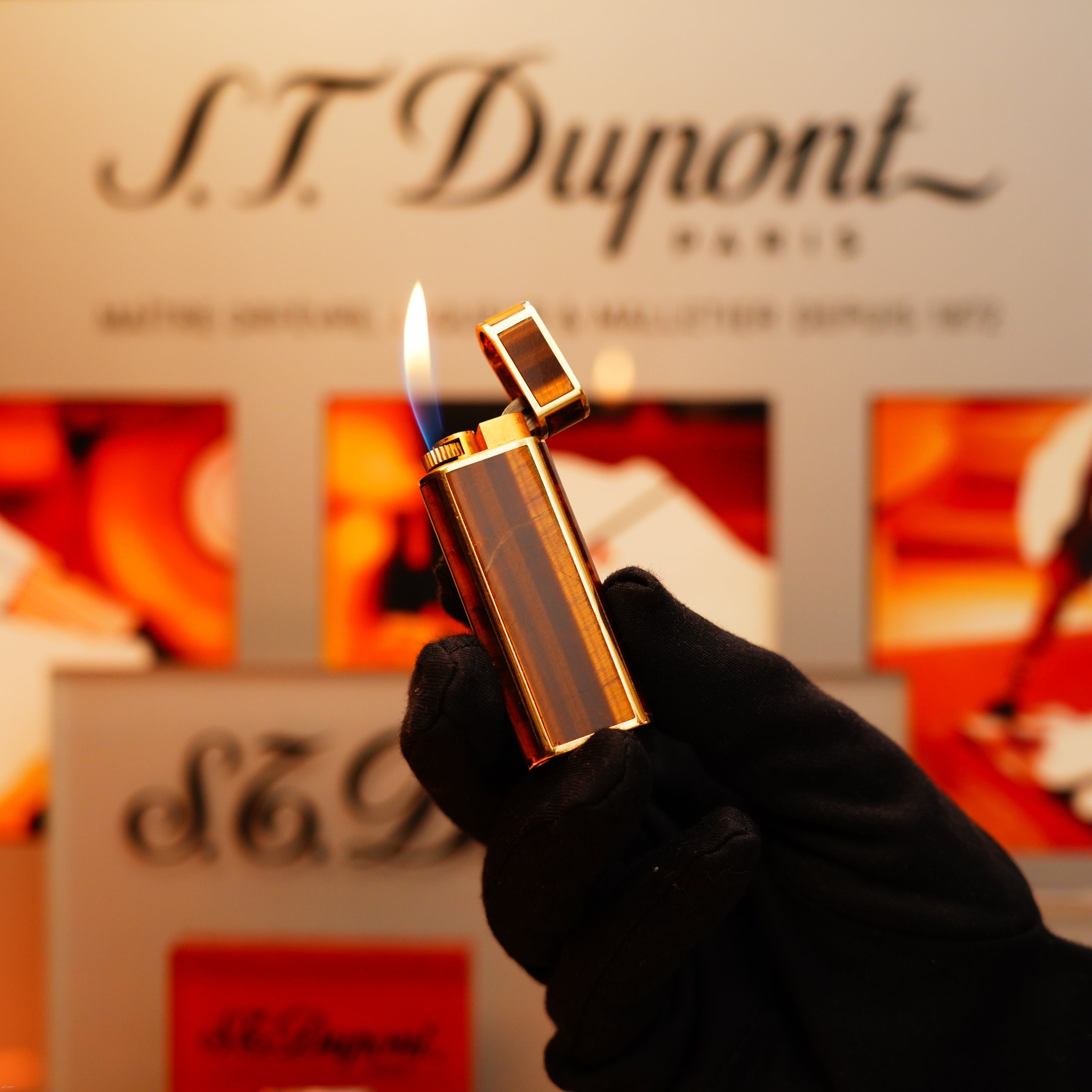 A gloved hand holds a lit Vintage 1970 Cartier Tiger's Eye Solid 18k Lighter with 750 hallmark by KirbyAllison.com, flame visible against a blurred branded background.