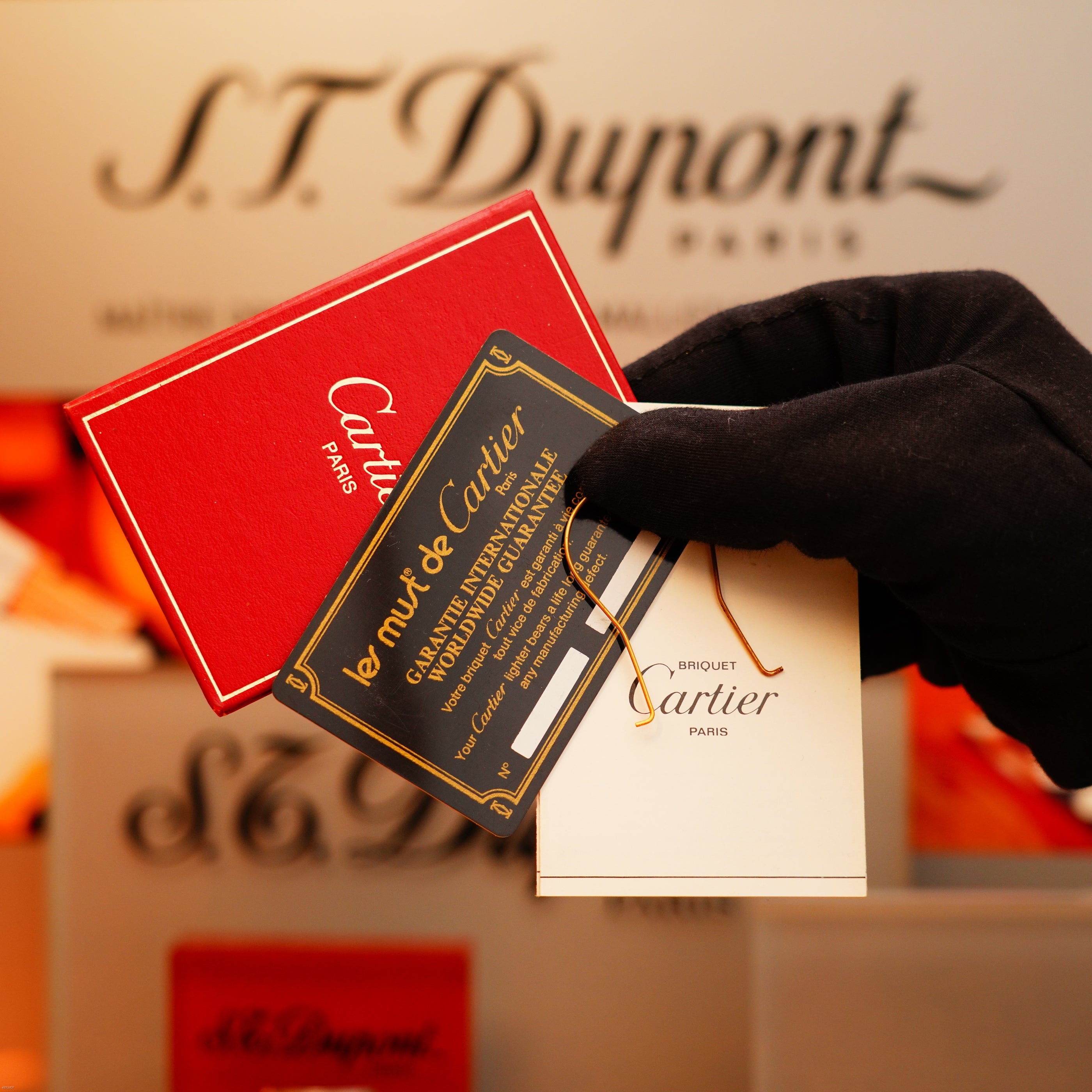 A gloved hand holds KirbyAllison.com product cards and packaging for the Vintage 1970 Cartier Extremely Rare Tiger's Eye Solid 18k Heavy Body Lighter with 750 hallmark, against a blurred branded background.