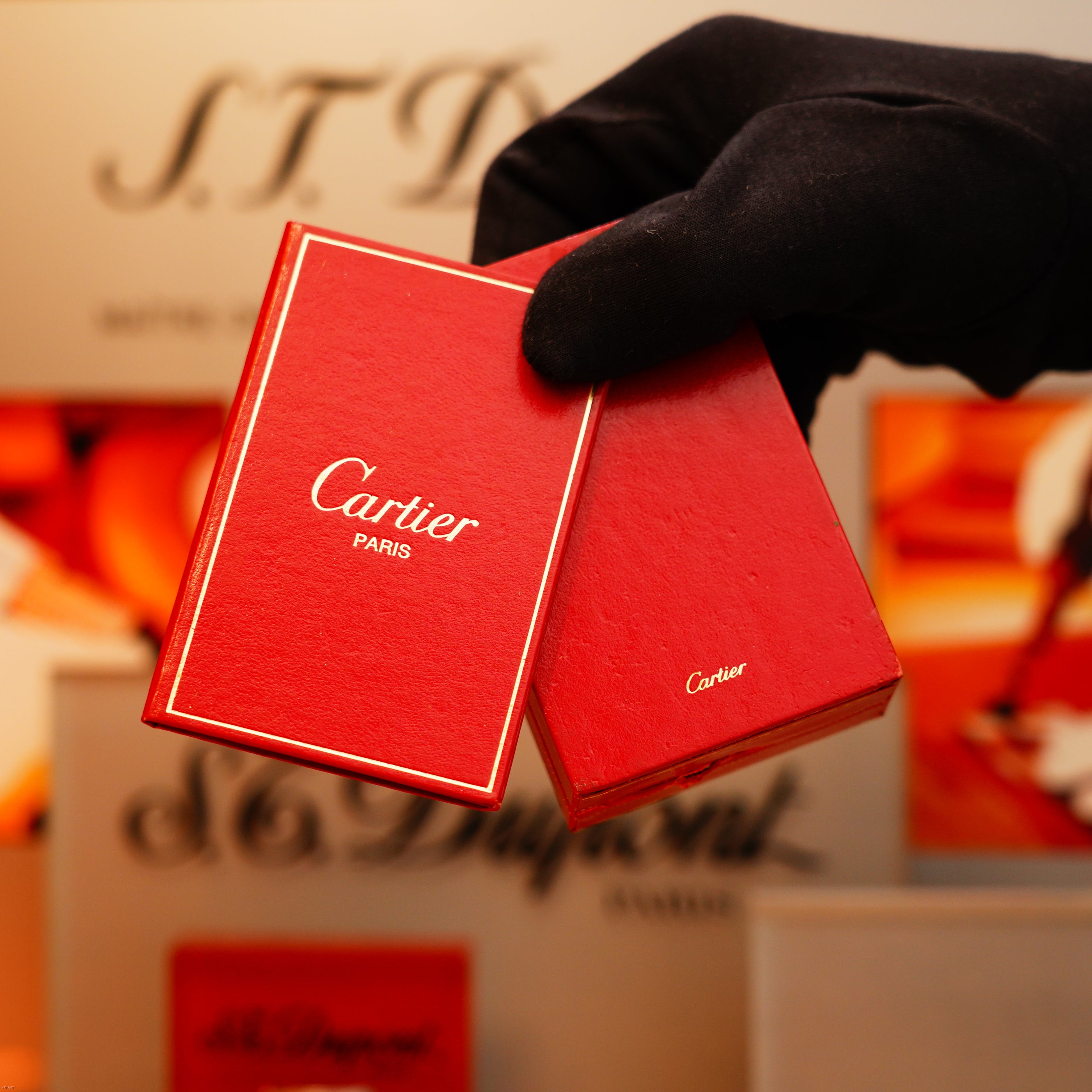 A gloved hand holds a Vintage 1970 Cartier Extremely Rare Tiger's Eye Solid 18k Lighter with 750 hallmark from KirbyAllison.com against a blurred luxury-branded background.
