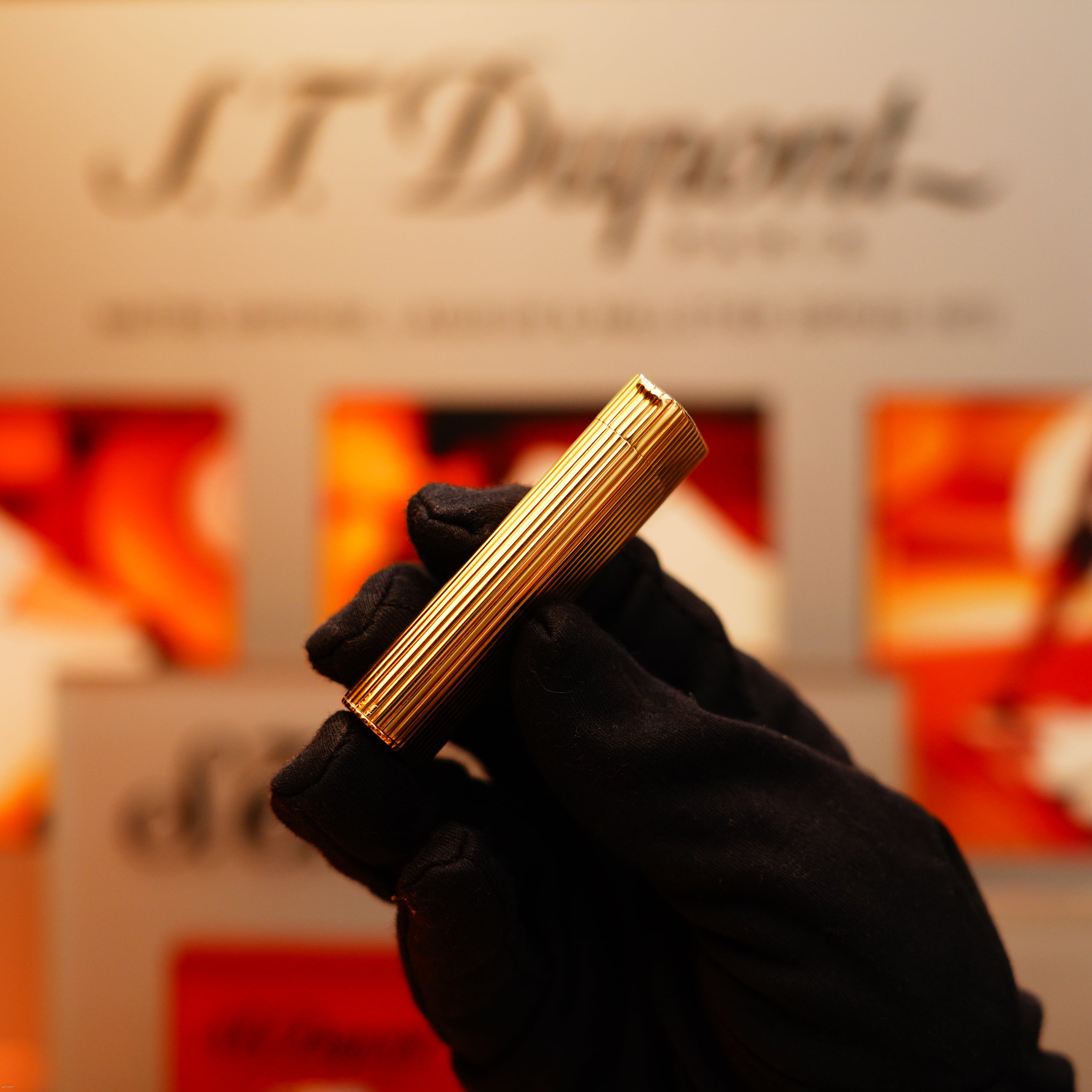 A gloved hand holds a Vintage 1970 Extremely Rare Cartier Solid 18k Gold Vertical Line Heavy Lighter with 750 hallmarks from KirbyAllison.com, set against a blurred branded backdrop.