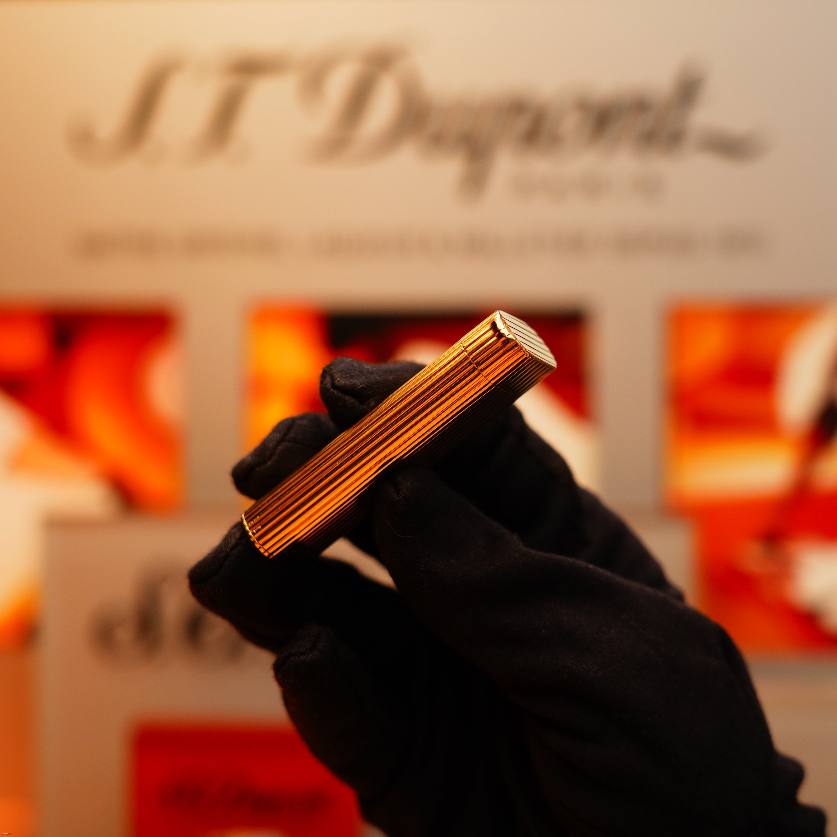 A hand in a black glove holds a Vintage 1970 Extremely Rare Cartier Solid 18k Gold Vertical Line Heavy Lighter with 750 hallmarks from KirbyAllison.com in front of a blurred S.T. Dupont Paris display.