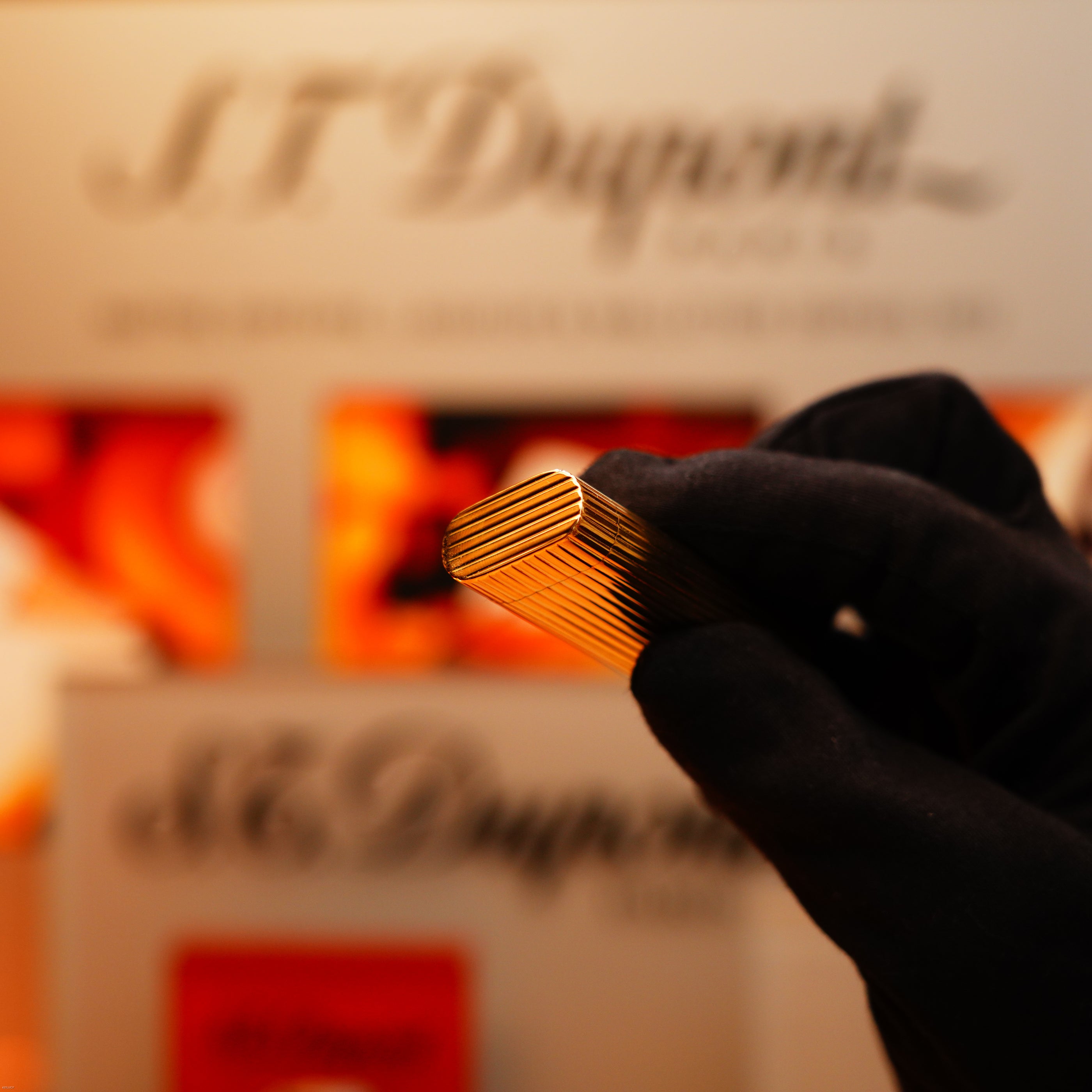 A gloved hand holds a Vintage 1970 Extremely Rare Cartier Solid 18k Gold Vertical Line Heavy Lighter with 750 hallmarks from KirbyAllison.com, with an S.T. Dupont display blurred in the background.