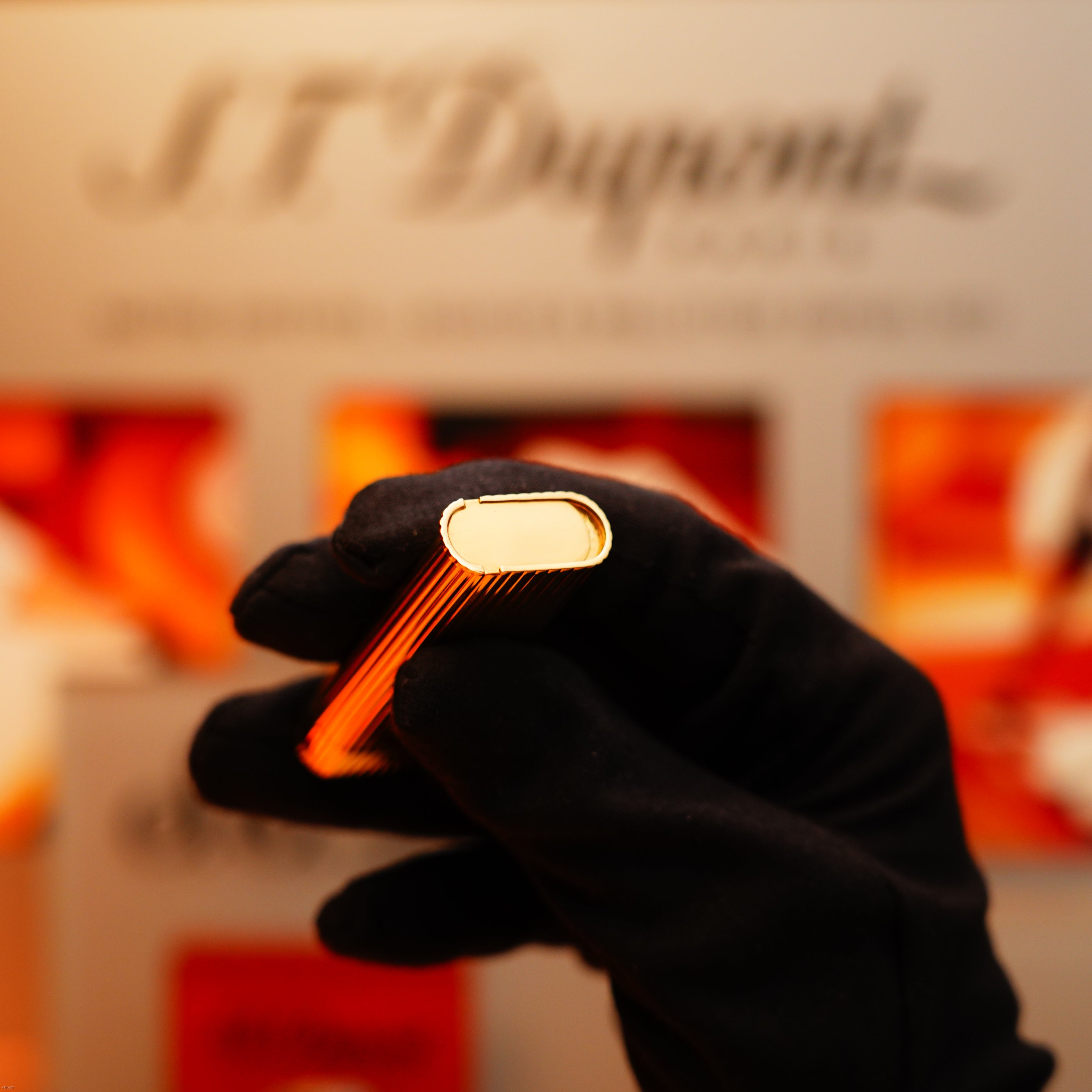 A gloved hand holds a Vintage 1970 Extremely Rare Cartier Solid 18k Gold Vertical Line Heavy with 750 hallmarks Lighter by KirbyAllison.com horizontally, with a blurred logo and display in the background.