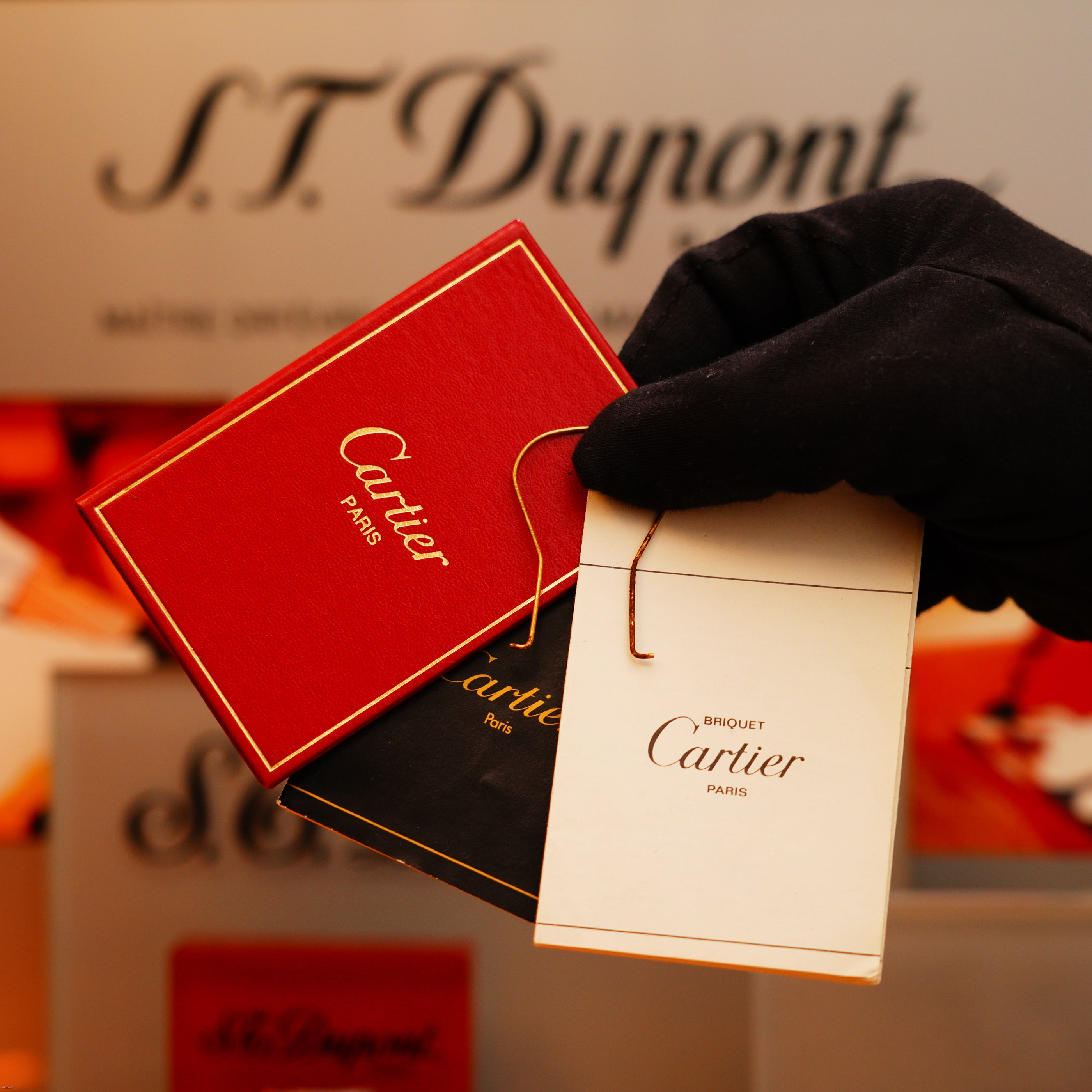 A gloved hand displays three Cartier cards in red, black, and white, with a Vintage 1970 Extremely Rare Cartier Solid 18k Gold Vertical Line Heavy Lighter from KirbyAllison.com seen blurred in the background.