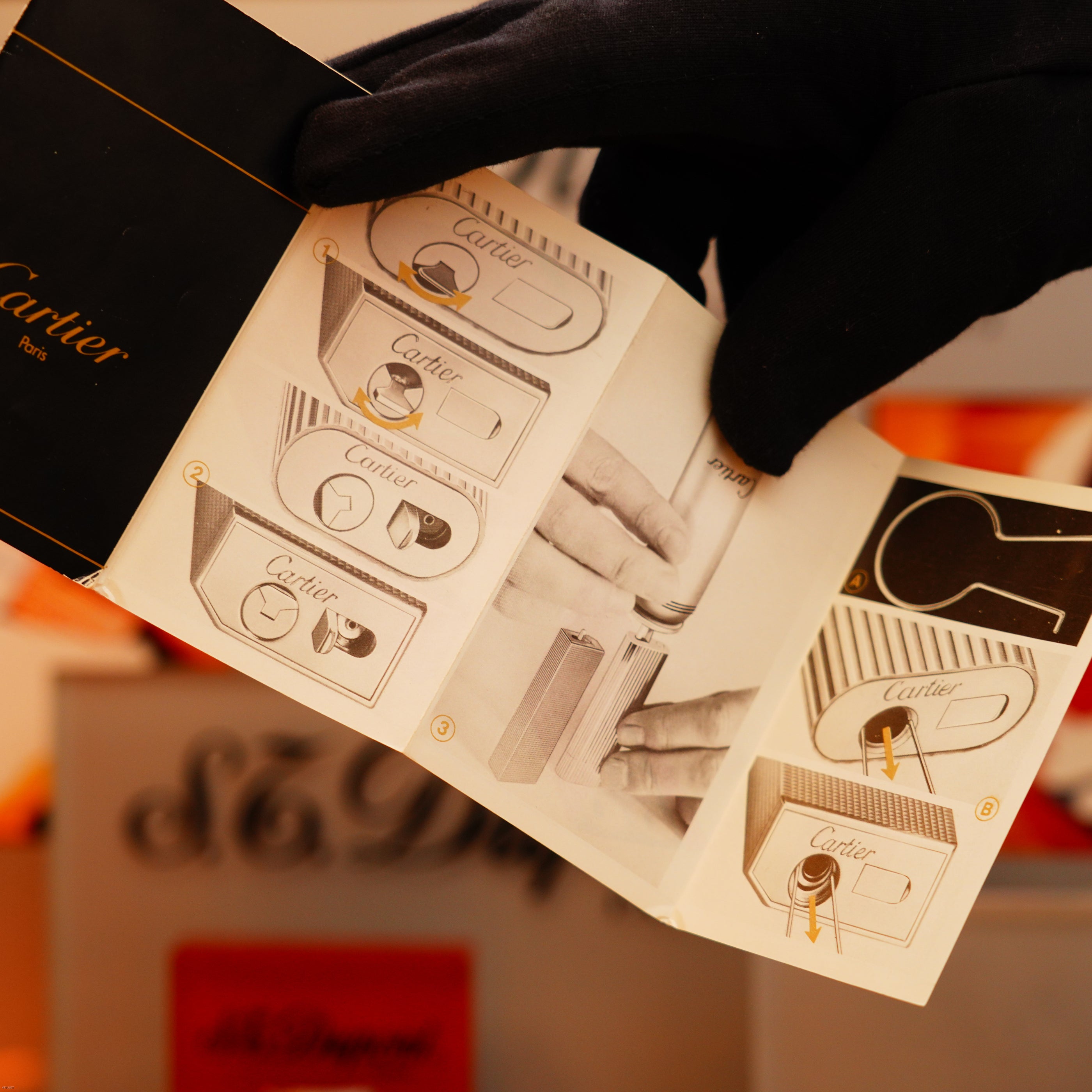 A gloved hand holds an open instruction manual with diagrams for the Vintage 1970 Extremely Rare Cartier Solid 18k Gold Vertical Line Lighter, offered by KirbyAllison.com.