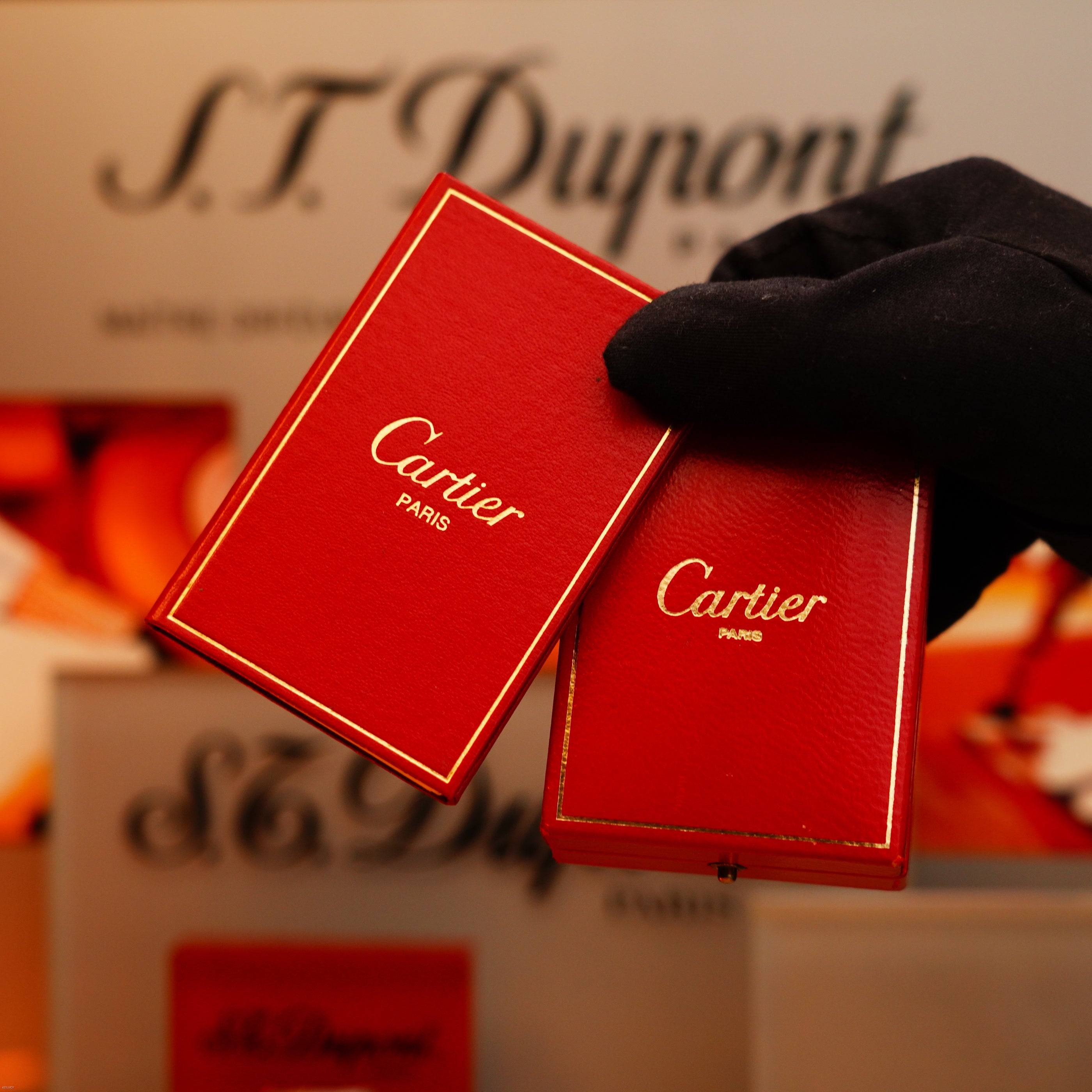 A gloved hand holds two red boxes with gold trim; in the background, "Vintage 1970 Extremely Rare Cartier Solid 18k Gold Vertical Line Heavy with 750 hallmarks Lighter" by KirbyAllison.com is visible.