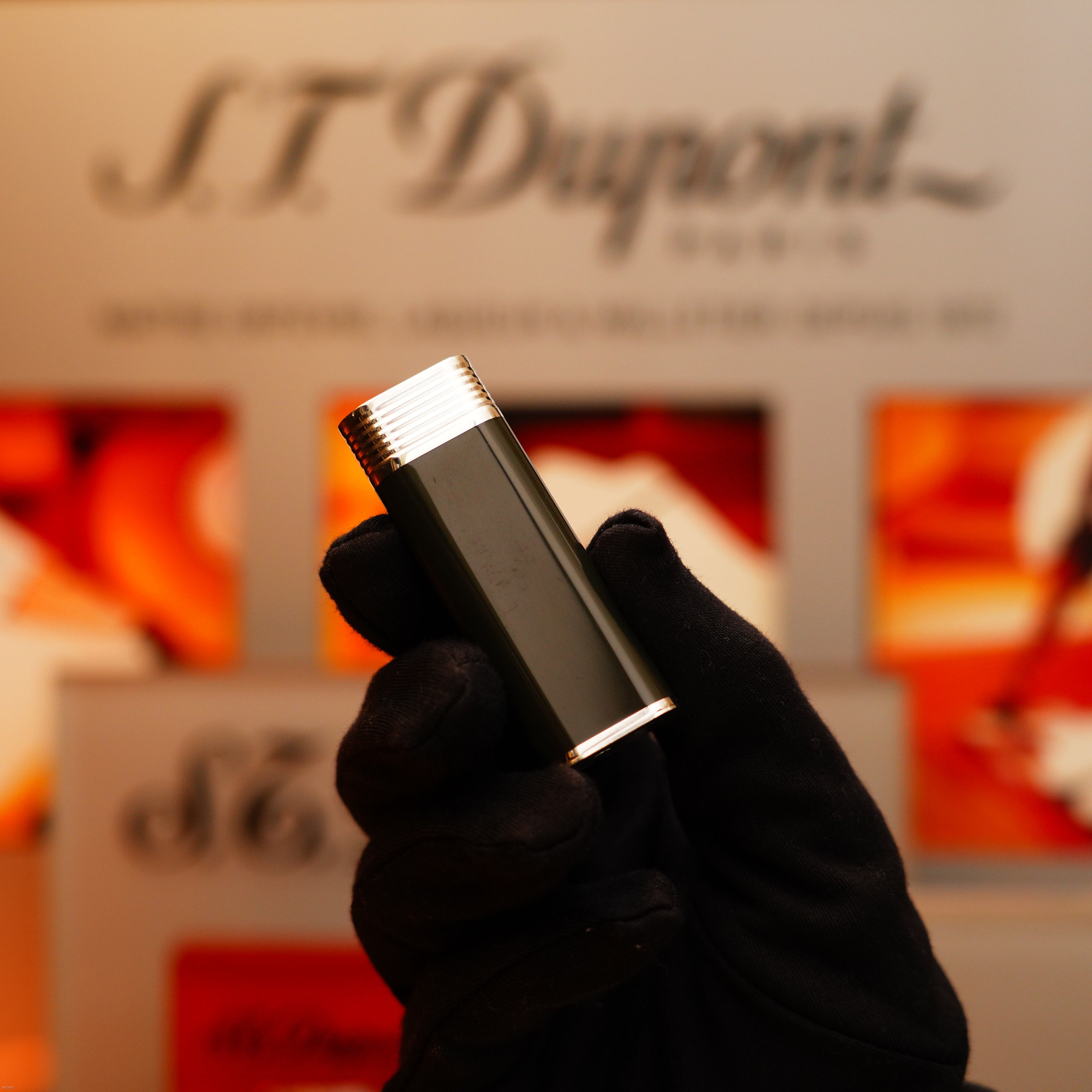 A gloved hand holds the Vintage 1970 Extremely Rare Cartier Solid 925 Sterling Silver Natural Full Dark Green Lacquer Rare Lighter from KirbyAllison.com, with a silver top, in front of a blurred S.T. Dupont display.