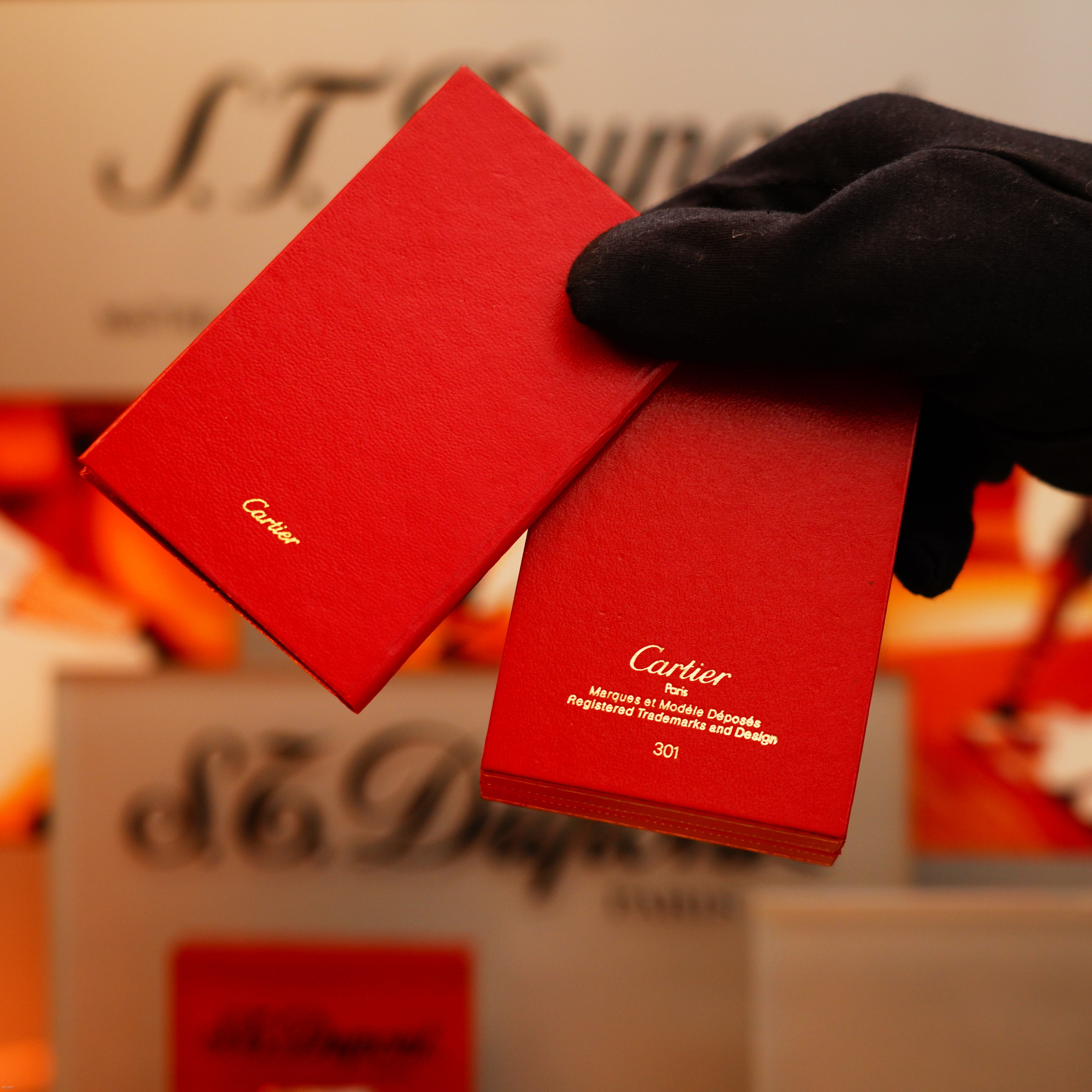 A gloved hand holds two red boxes against a blurred background with KirbyAllison.com branding, showcasing the Vintage 1970 Extremely Rare Cartier Solid 925 Sterling Silver Dark Green Lacquer Lighter.