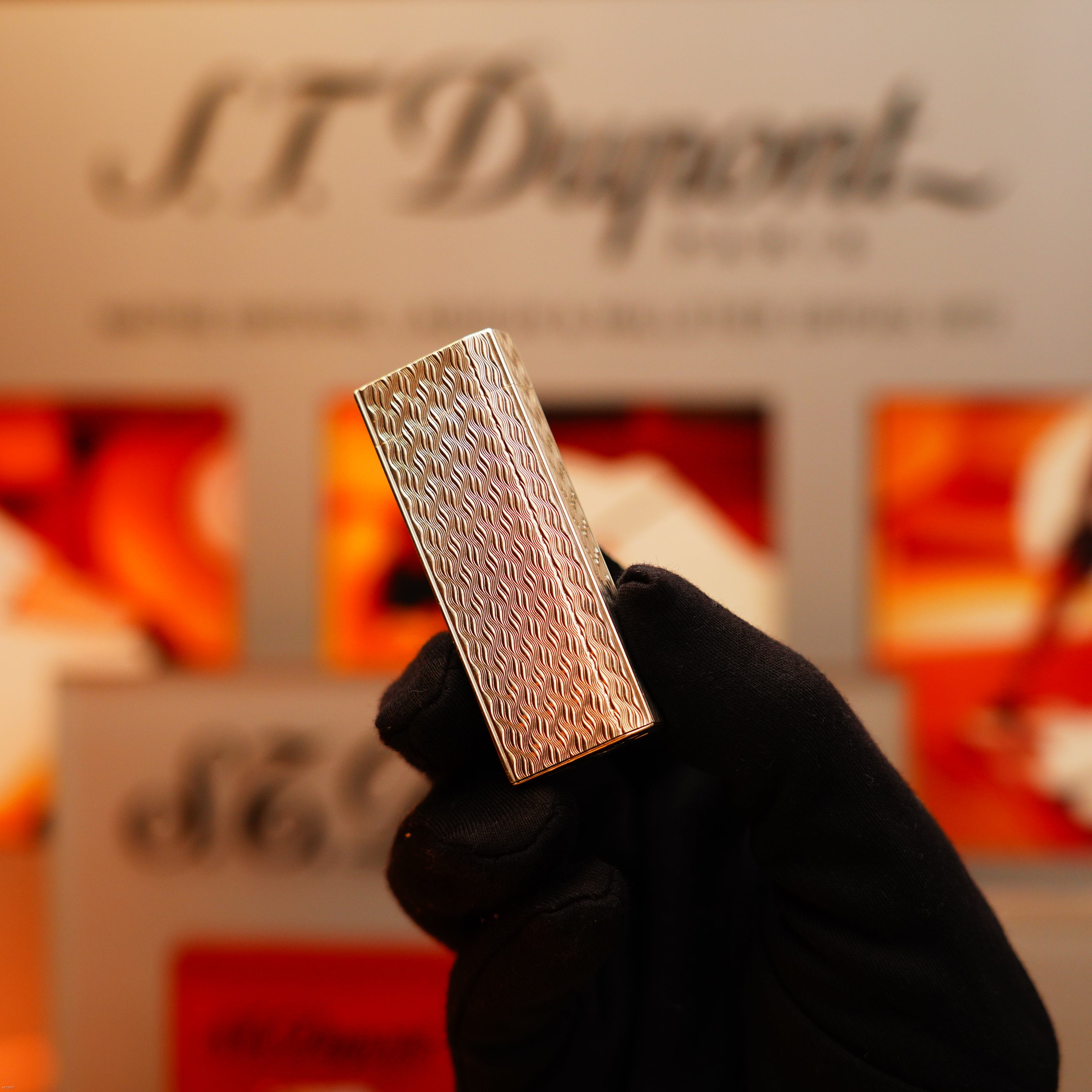 A gloved hand holds a Vintage 1970 Early Cartier Production Pentagone Solid 925 Extremely Heavy Sterling Silver Lighter by Cartier in front of a blurred branded background.