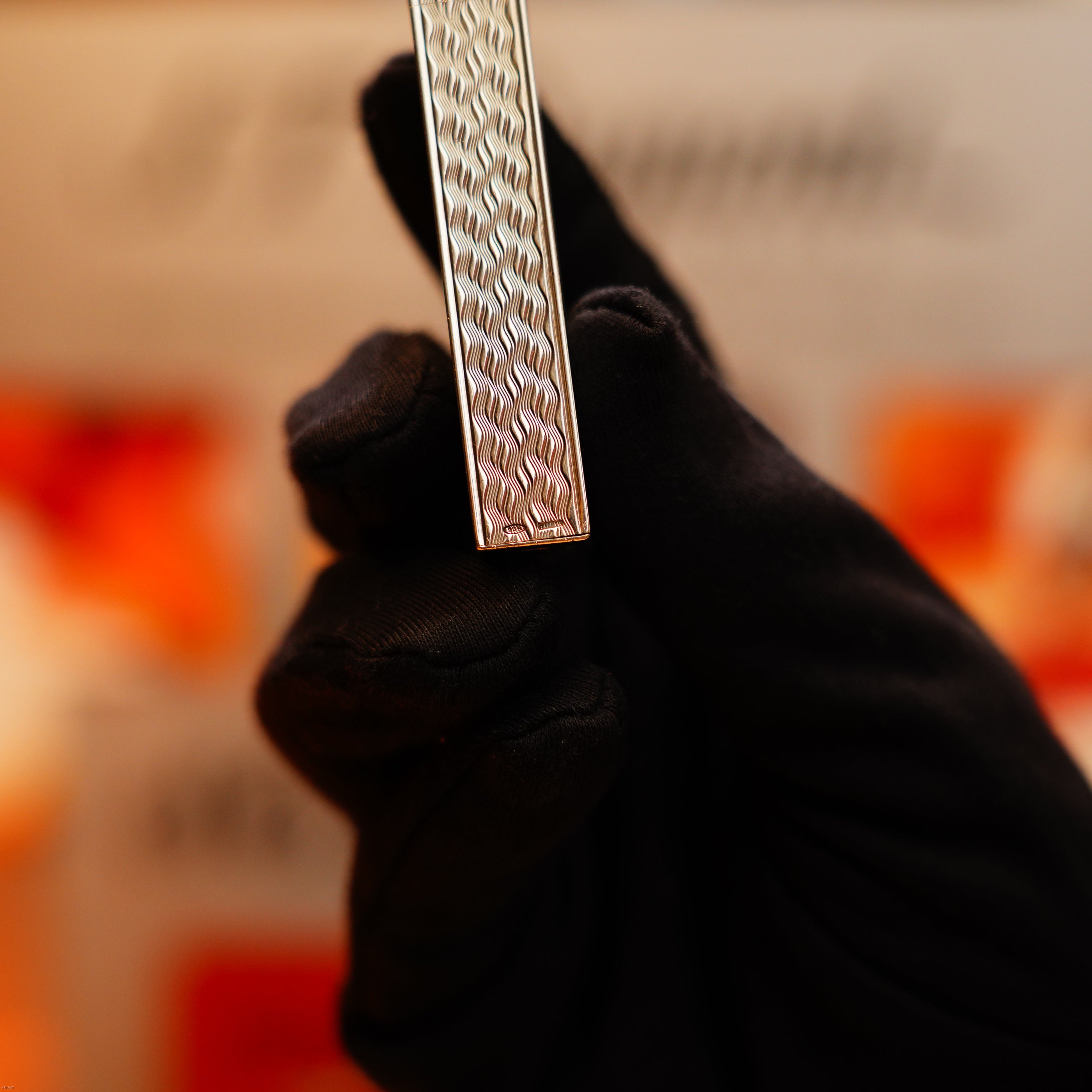 A gloved hand holds a Vintage 1970 Early Cartier Pentagone Solid 925 Heavy Sterling Silver Lighter against a blurred, warm-toned background. The lighter features Cartier’s signature textured, patterned metallic surface.