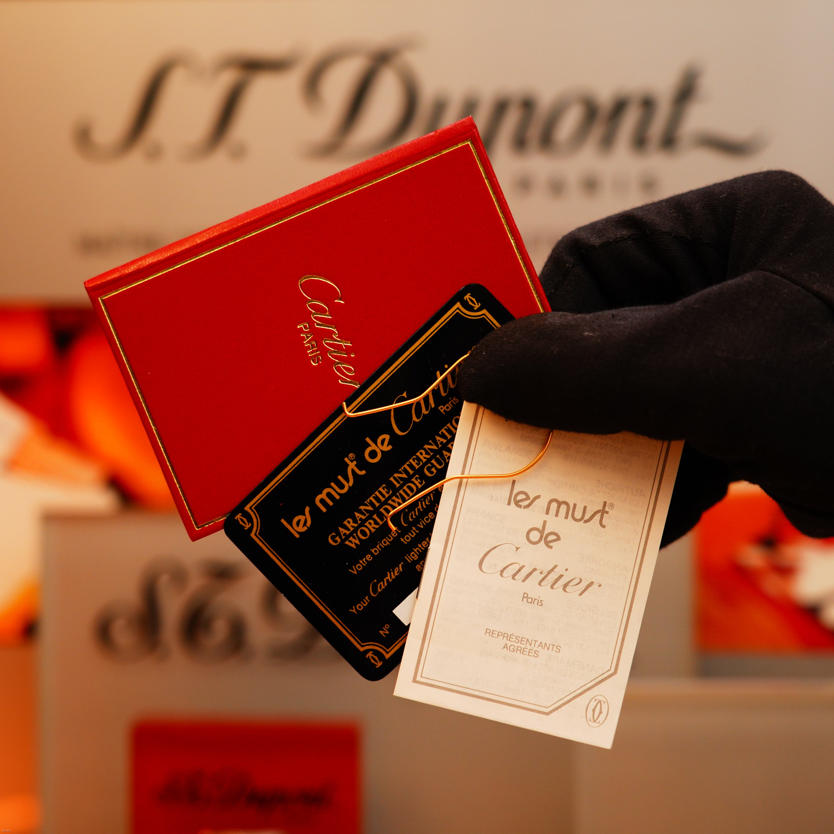 A gloved hand holds red and white Cartier cards with gold and black text, displaying the Vintage 1970 Early Cartier Production Pentagone Solid 925 Heavy Sterling Silver Lighter; an ST Dupont sign is blurred in the background.