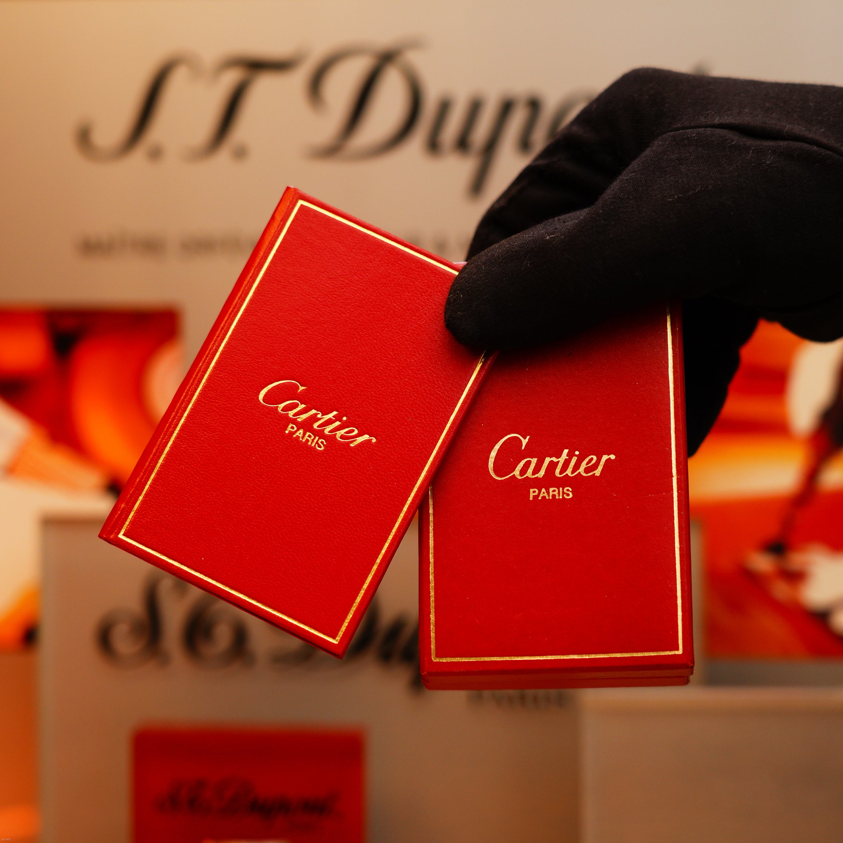 A gloved hand holds two red Cartier boxes with gold trim in front of a blurred S.T. Dupont display, featuring the Vintage 1970 Early Cartier Production Pentagone Solid 925 Extremely Heavy Sterling Silver Lighter.